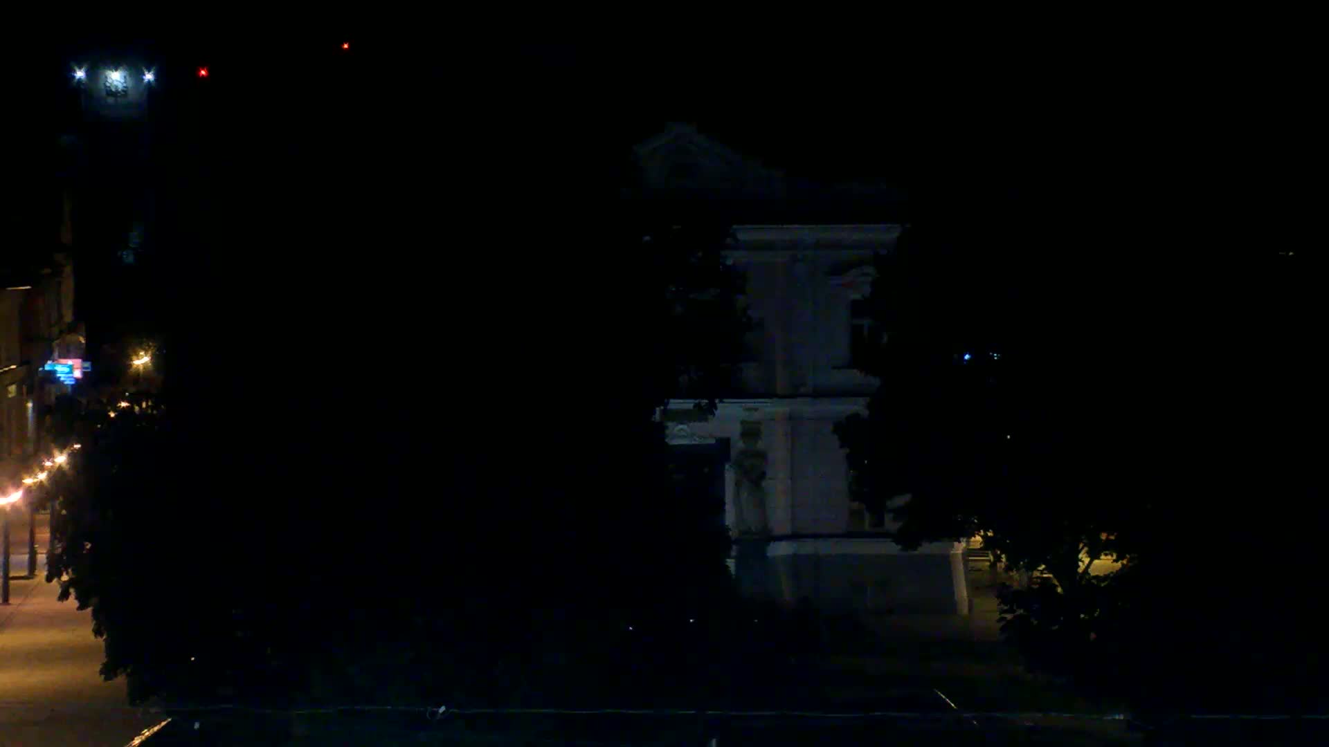 A dimly lit nighttime street scene features a large building partially obscured by trees, with faint streetlights visible in the distance.
