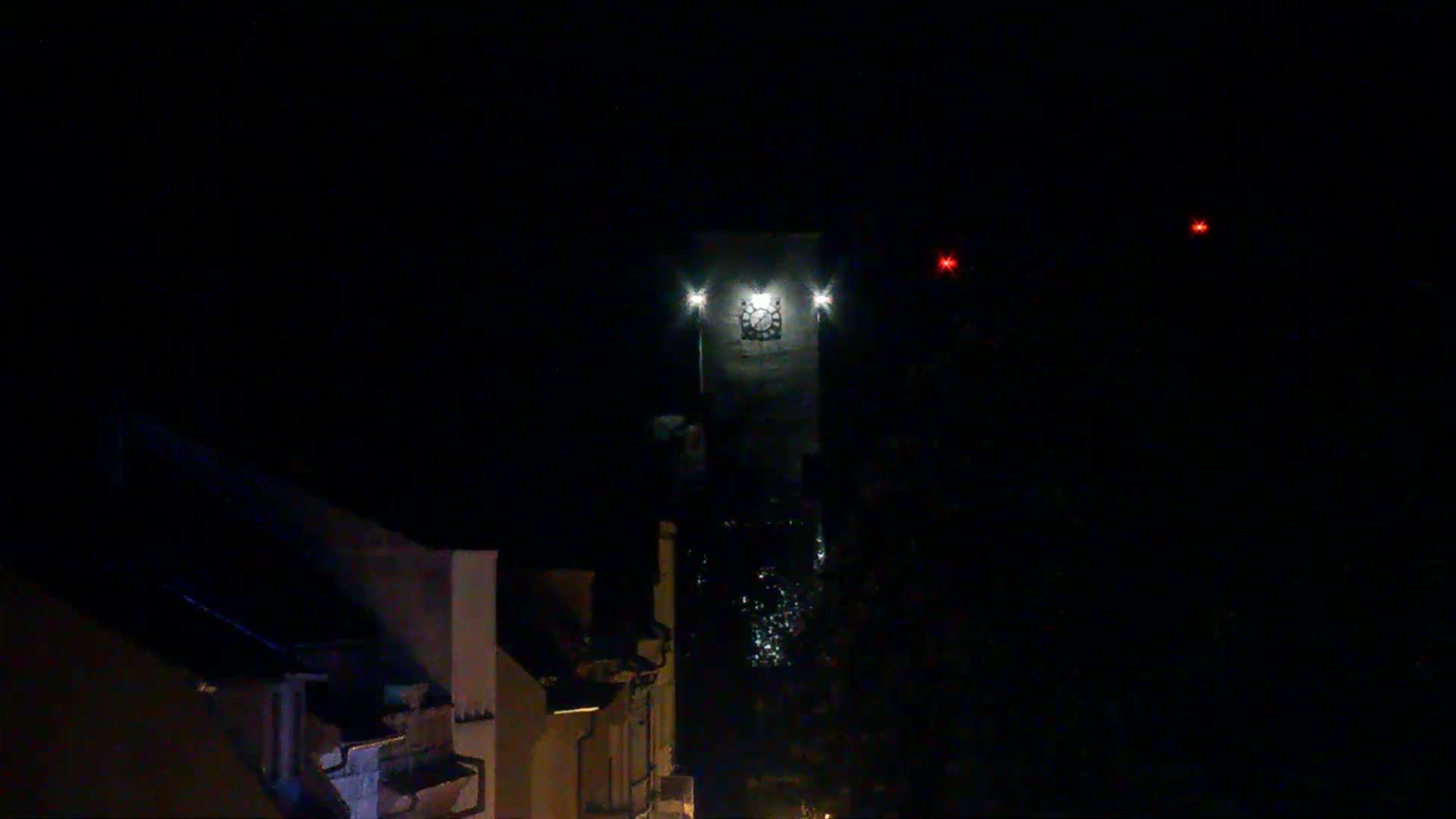 A tall, illuminated clock tower stands in the darkness of night, with faint lights visible below and distant red lights in the sky.