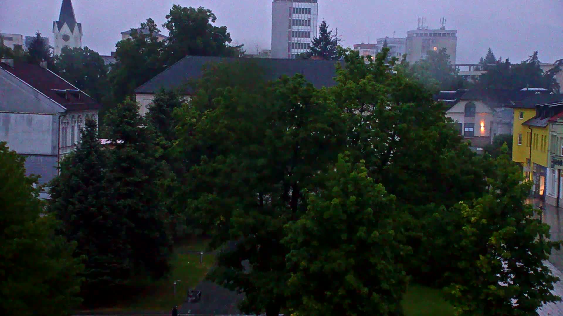 A rainy overcast day reveals a town square surrounded by lush green trees and buildings, including a church steeple and multi-story structures.