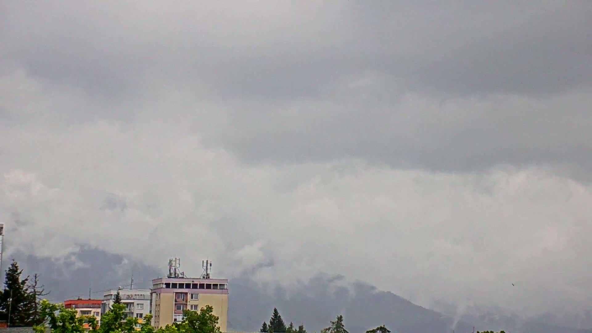 A cloudy, overcast day reveals a cityscape with several buildings, including a tall apartment building, set against a backdrop of hazy mountains.