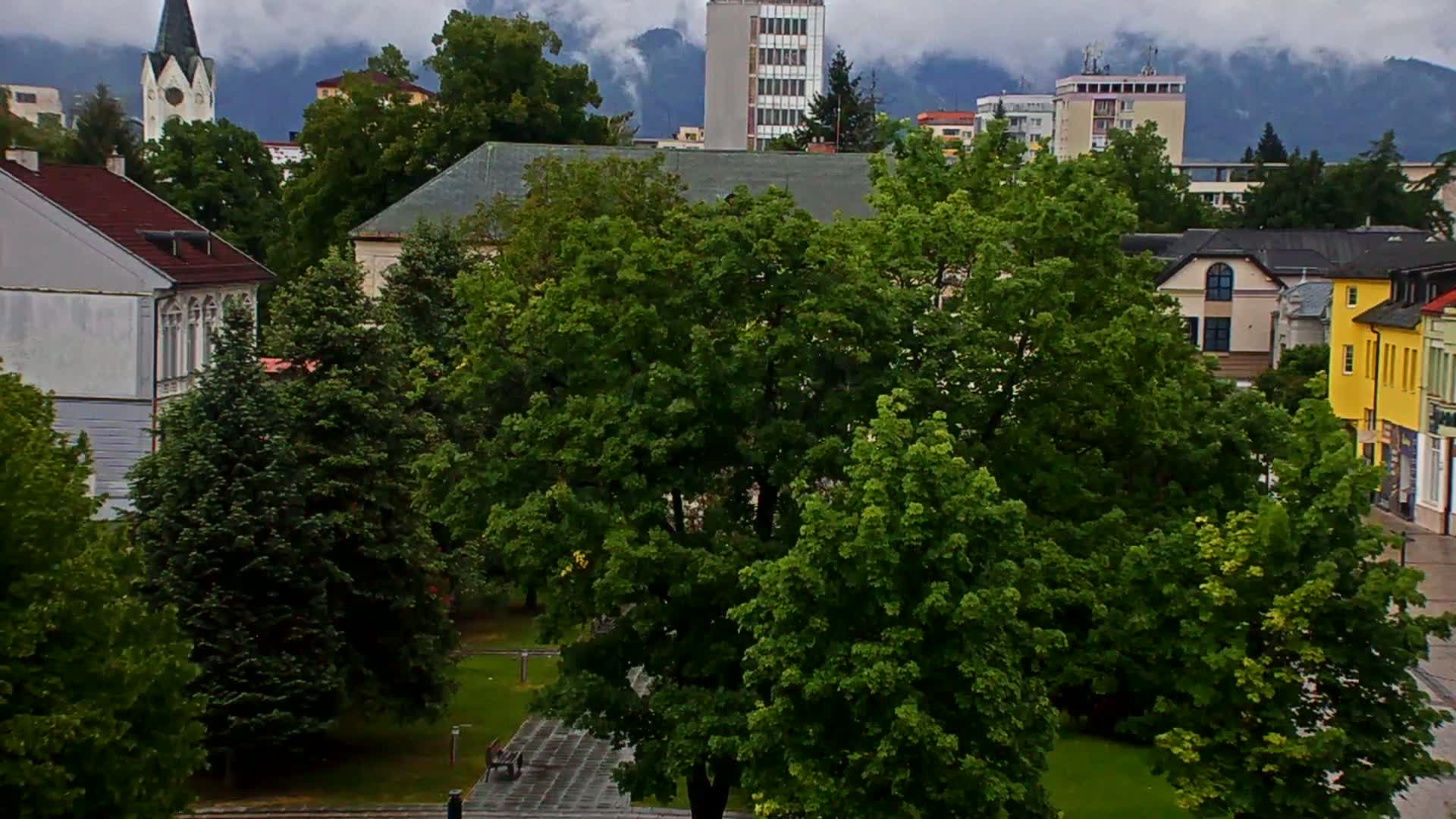 A town square with lush green trees, surrounded by buildings of varying heights and architectural styles, under a cloudy sky.