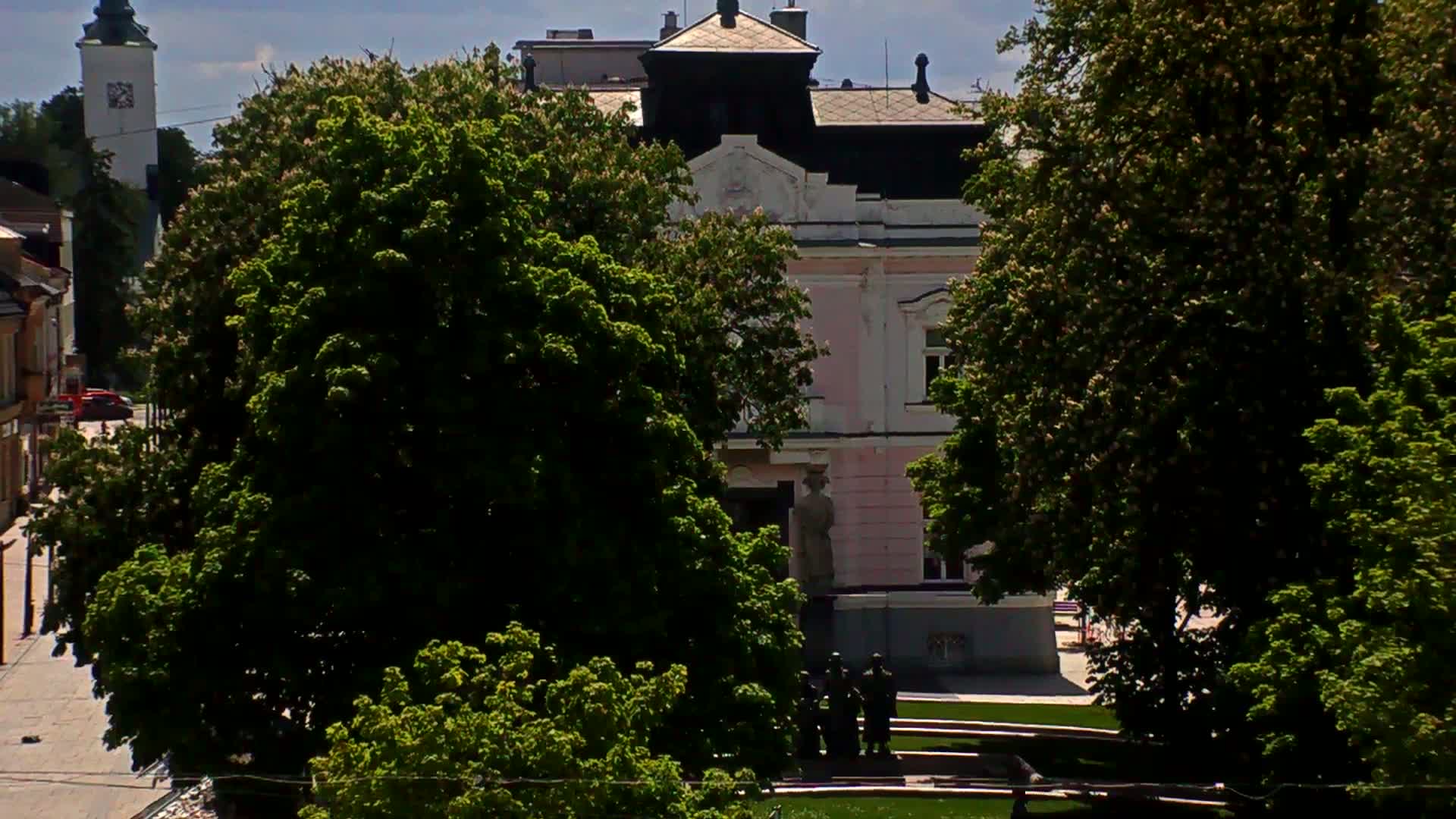 A pink building with a dark roof is partially obscured by lush green trees on a sunny day.