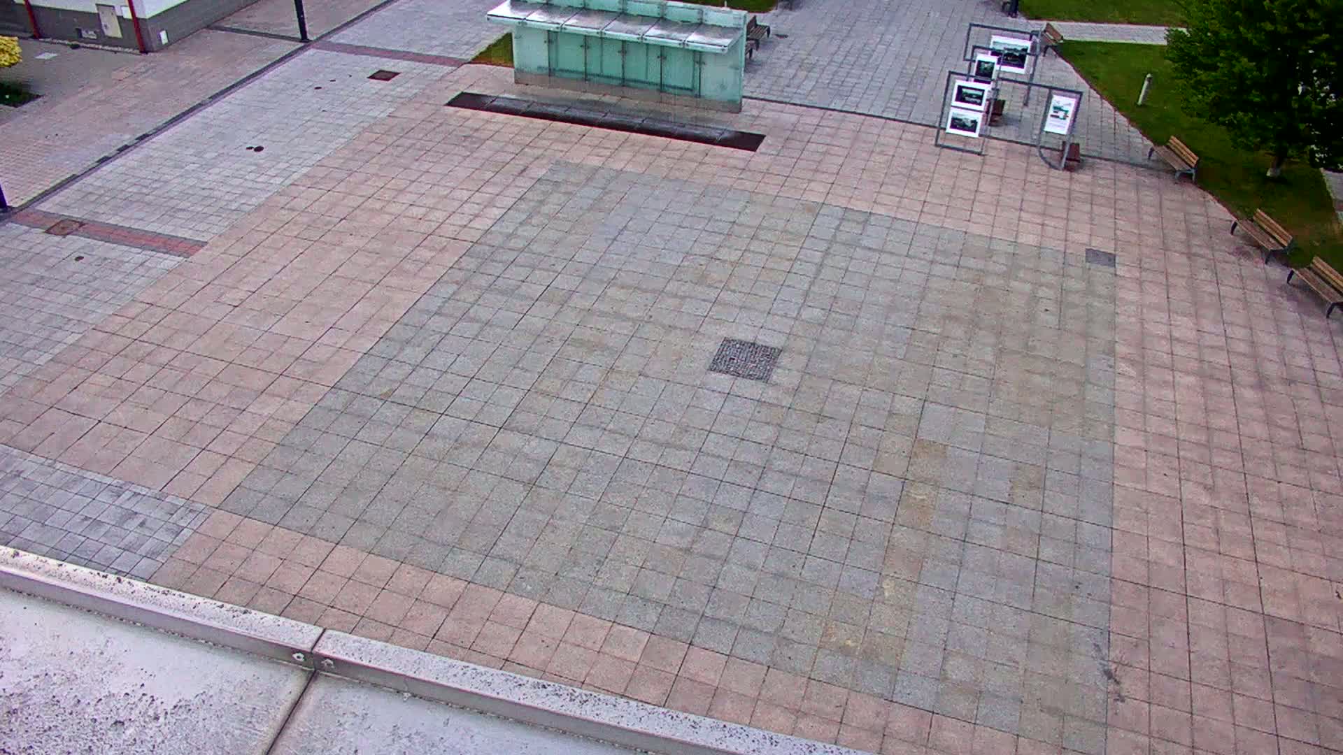 An overhead view shows a large paved plaza with a smaller, darker-colored square within it, bordered by lighter-colored paving stones, alongside a small glass kiosk and several park benches.