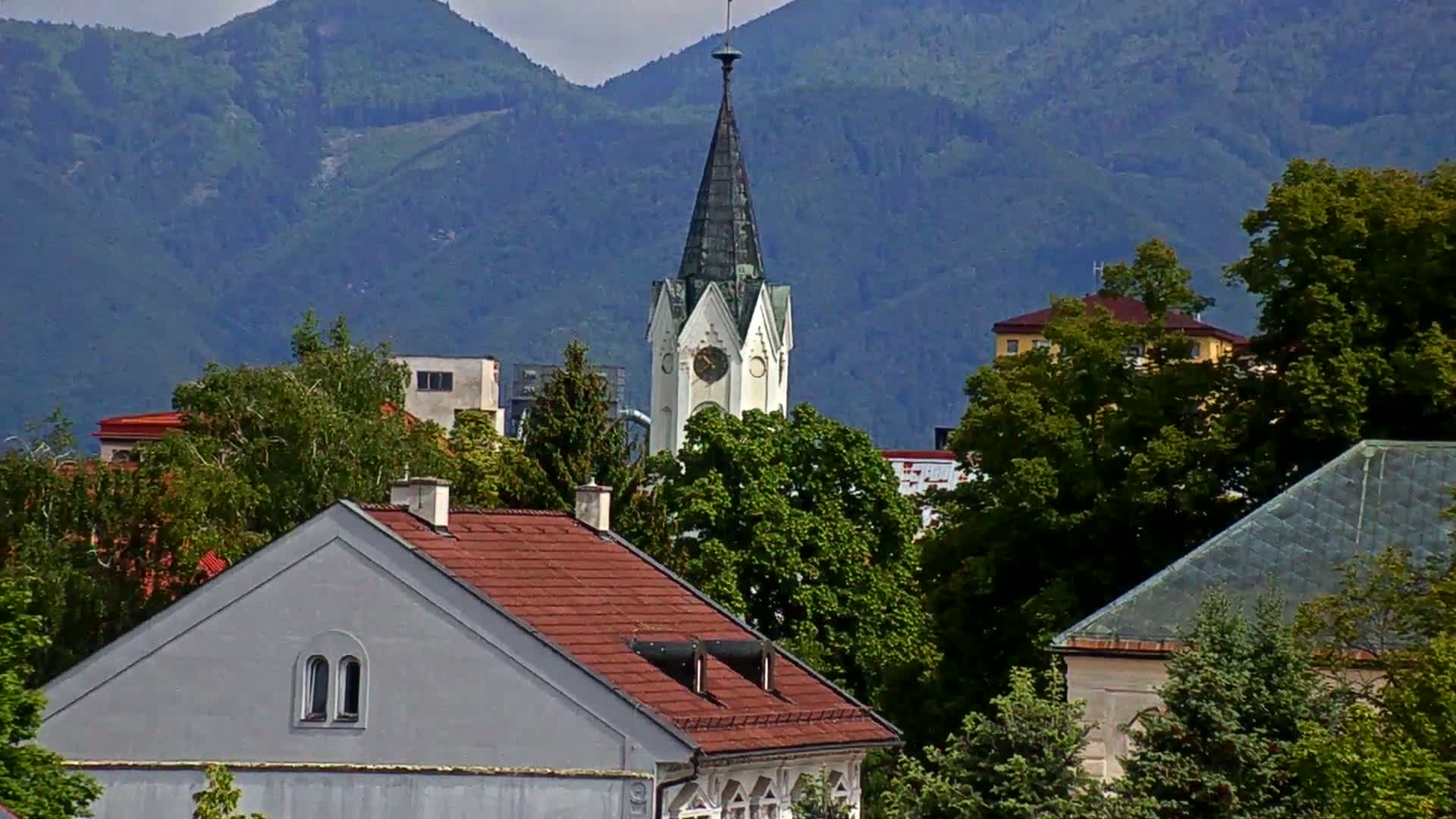 A church steeple rises above a collection of houses and lush green trees against a backdrop of hazy blue mountains.