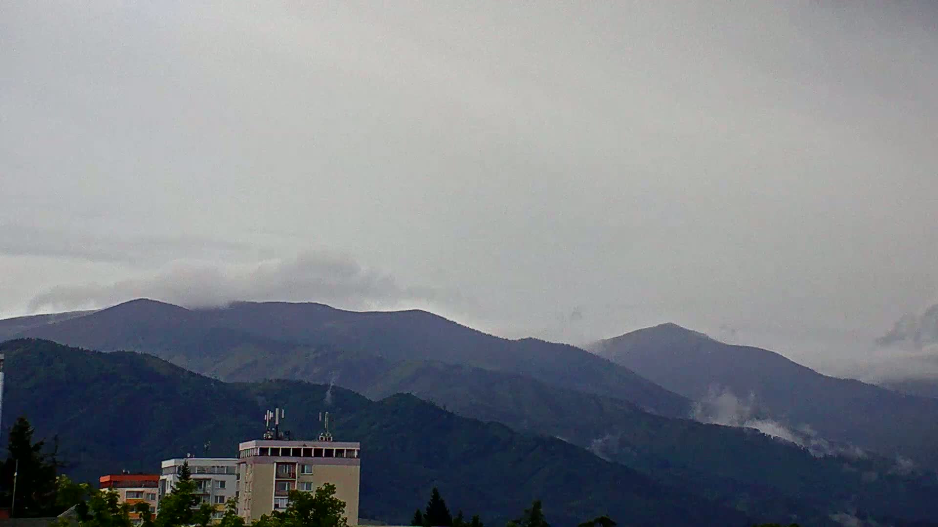 A cloudy, overcast day reveals a view of dark green mountains partially shrouded in mist, with several buildings visible in the foreground.