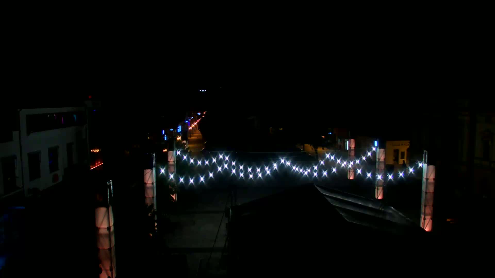 A nighttime street scene shows a string of lights strung across a roadway between illuminated columns, with buildings and streetlights visible on either side.