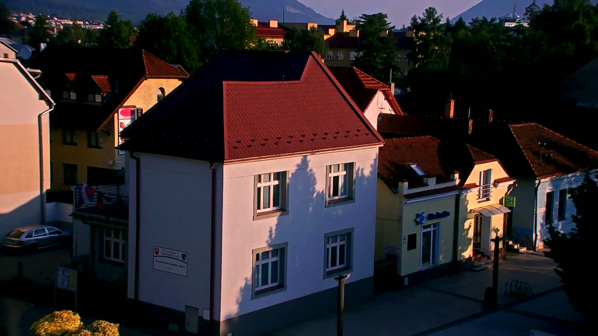 A light-grey two-story building with a reddish-brown roof sits among other houses in a town at sunset or sunrise, with shadows cast by trees on the building's facade.