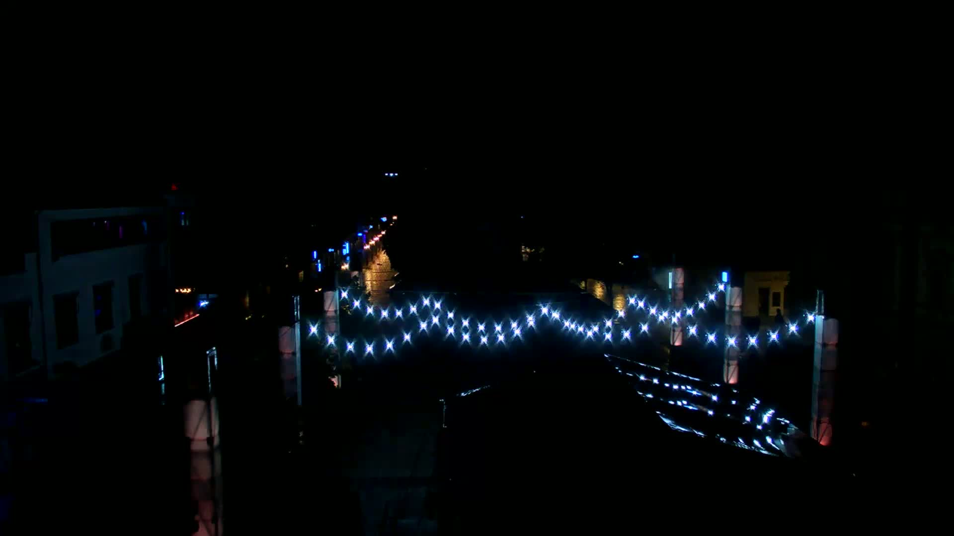 A nighttime view of a street with buildings on either side, illuminated by string lights strung across the road.