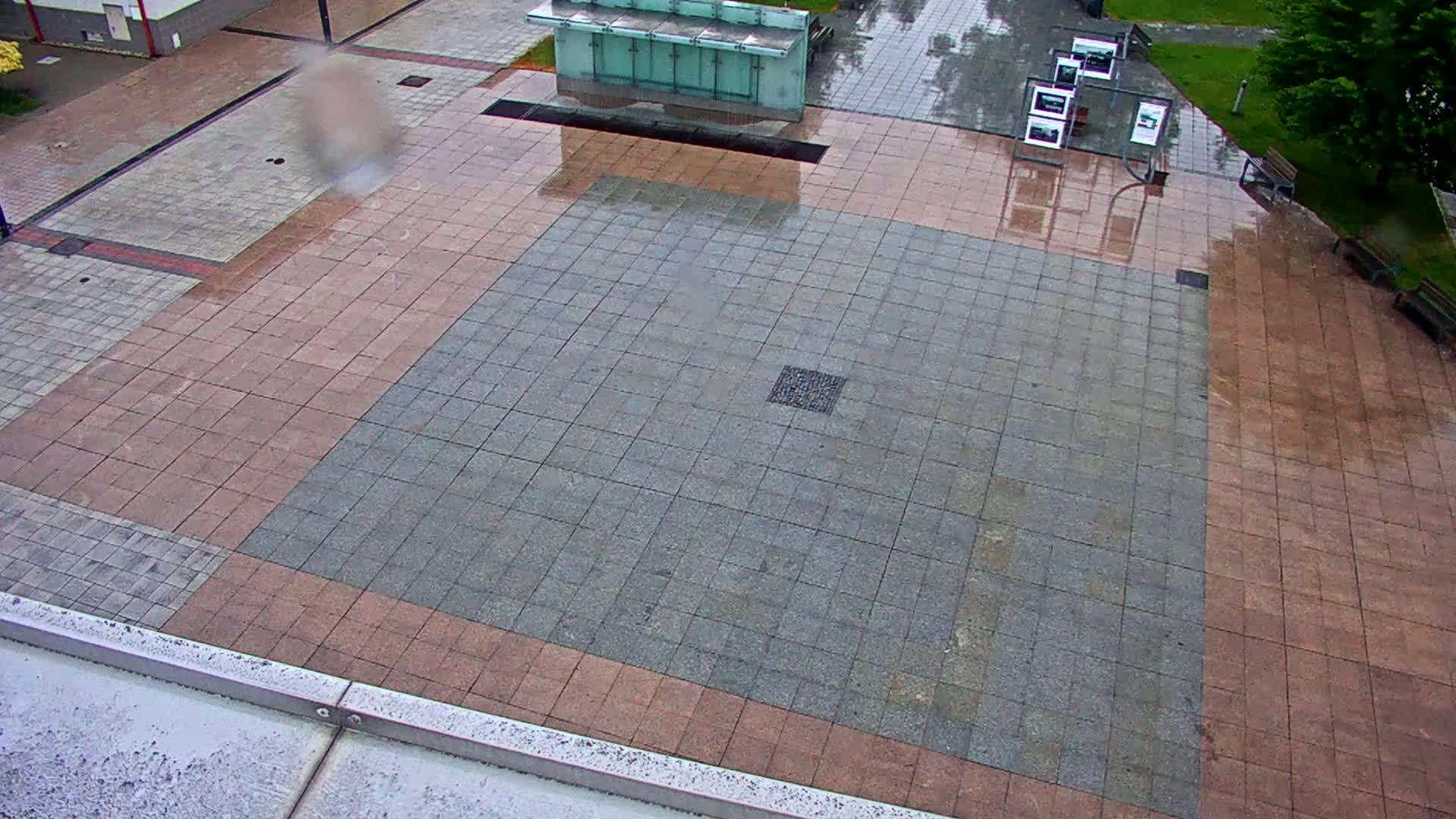 A wet, paved plaza with a large, square section of gray tiles bordered by reddish-brown tiles.