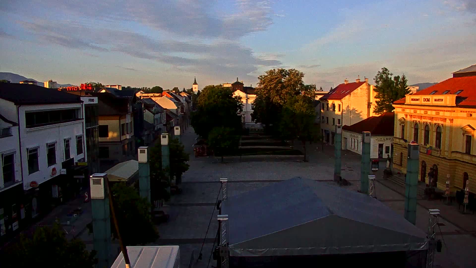 A town square at sunrise, with several buildings lining the edges, and a large covered stage in the foreground.