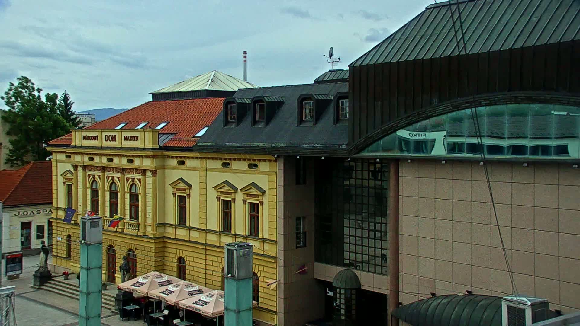 A yellow building with a red-tiled roof stands adjacent to a modern, tan building with a large arched window reflecting another structure, all under a cloudy sky.