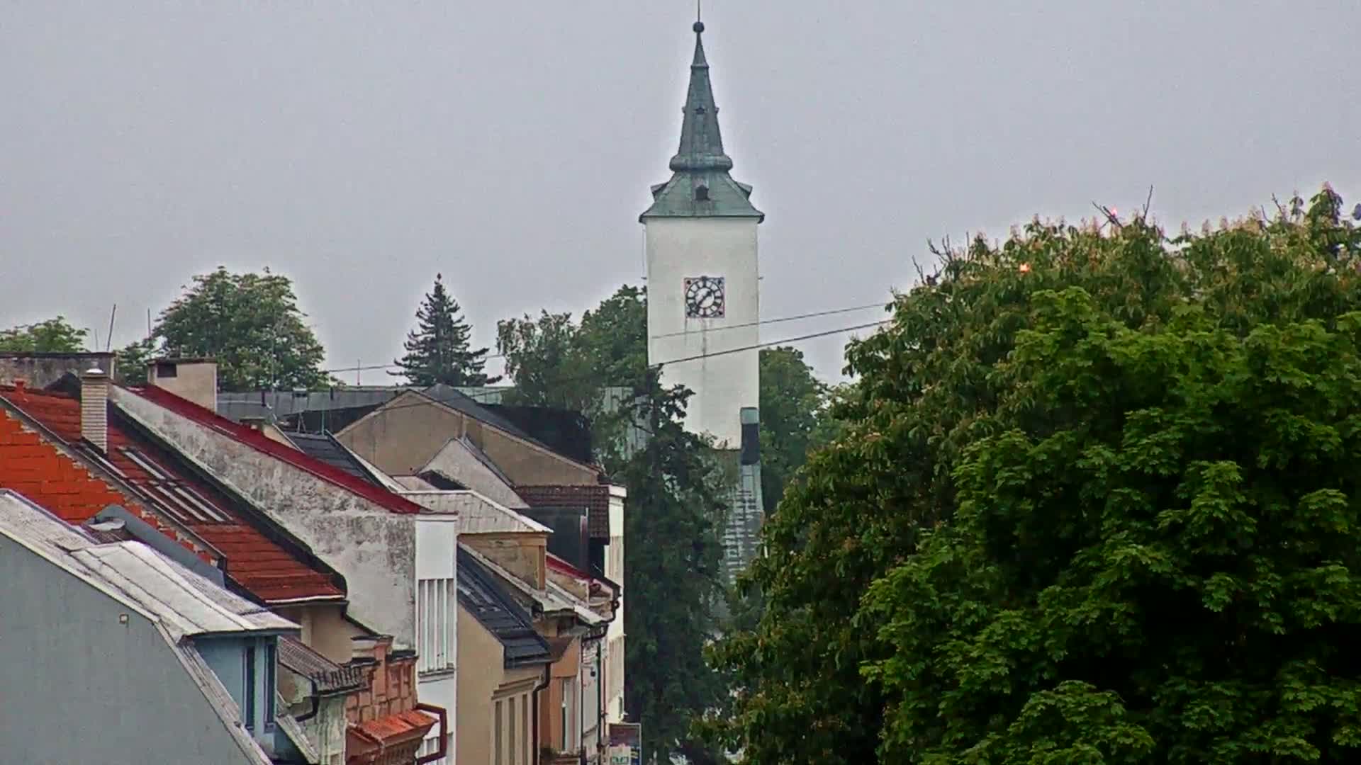 A white clock tower rises above a row of houses with red-tiled roofs under an overcast sky.