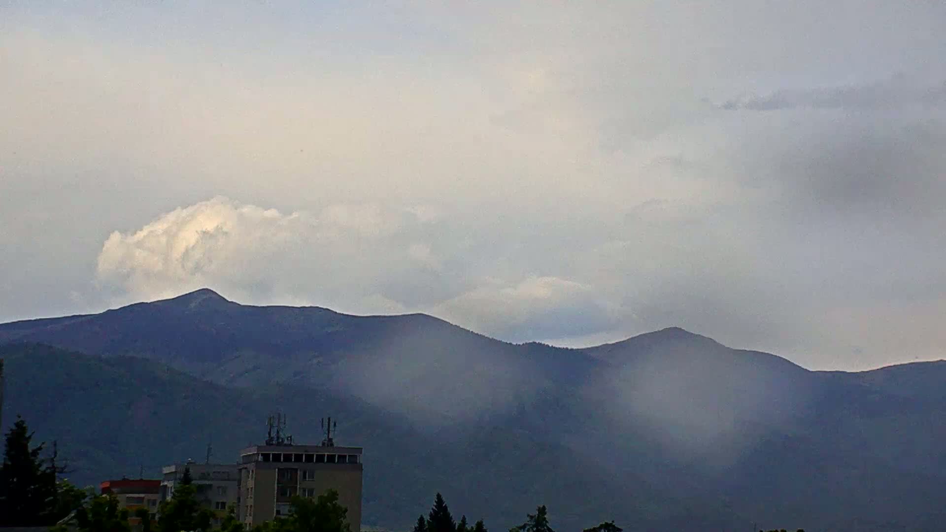 A partly cloudy sky hangs over a hazy mountain range, partially obscuring a city with a multi-story building in the foreground.