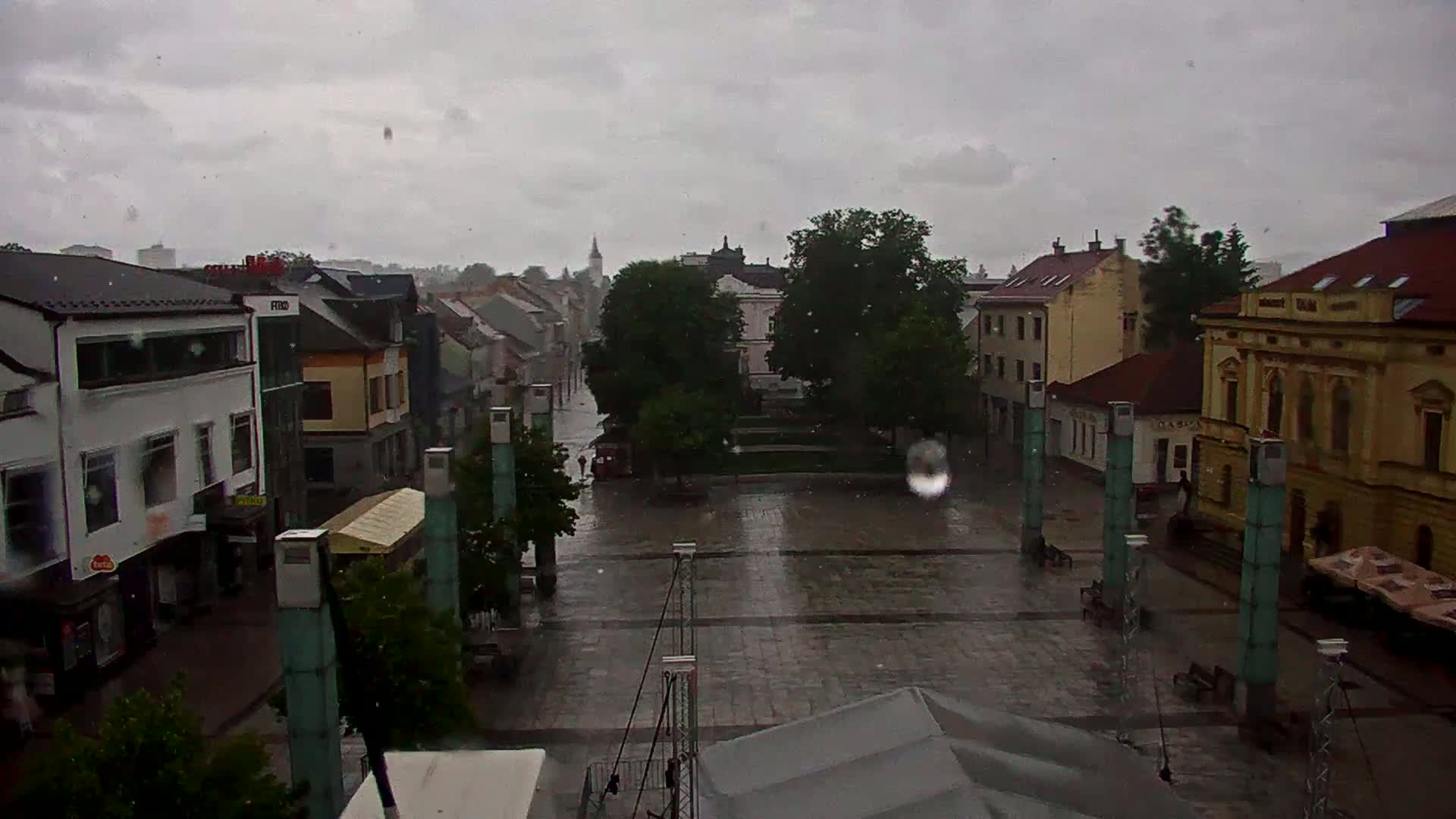A rain-soaked town square, surrounded by buildings, with several people walking in the distance under a gray, overcast sky.