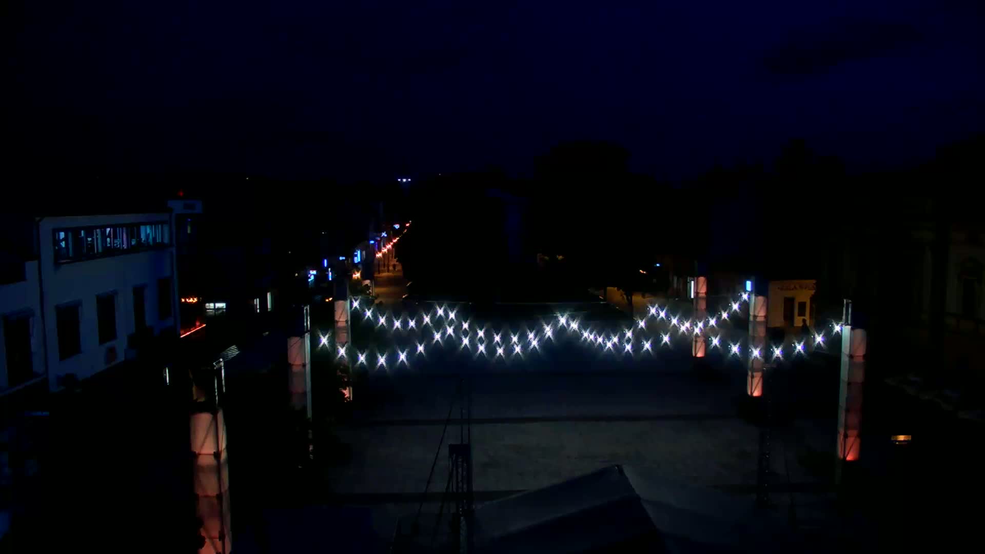 A nighttime view of a town square illuminated by strings of lights strung between tall posts, with buildings lining the street in the background under a dark sky.