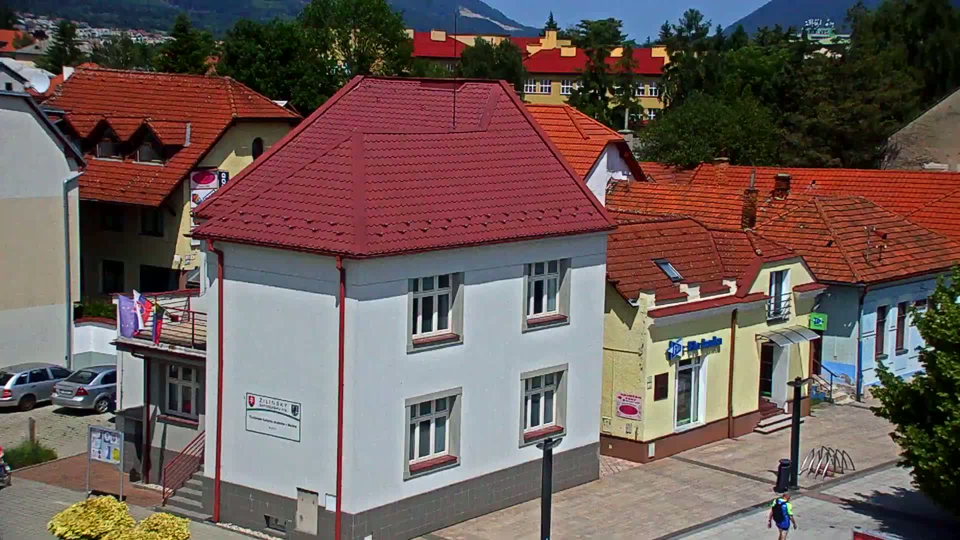 A white building with a red roof sits among other buildings with red-tiled roofs, under a sunny sky, in a town nestled against mountains.