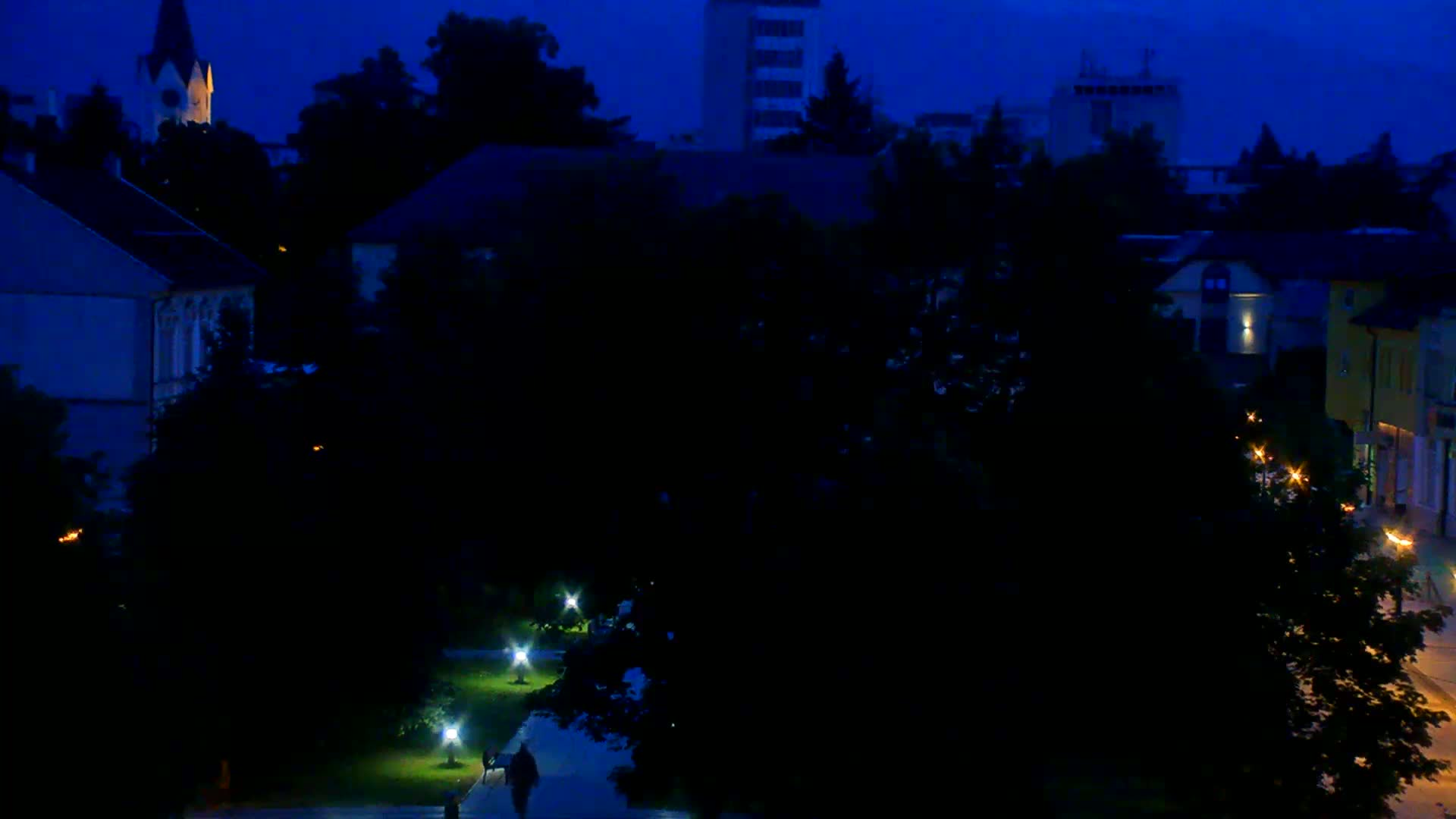 A nighttime view of a town square, with dimly lit buildings and trees, under a dark blue twilight sky.