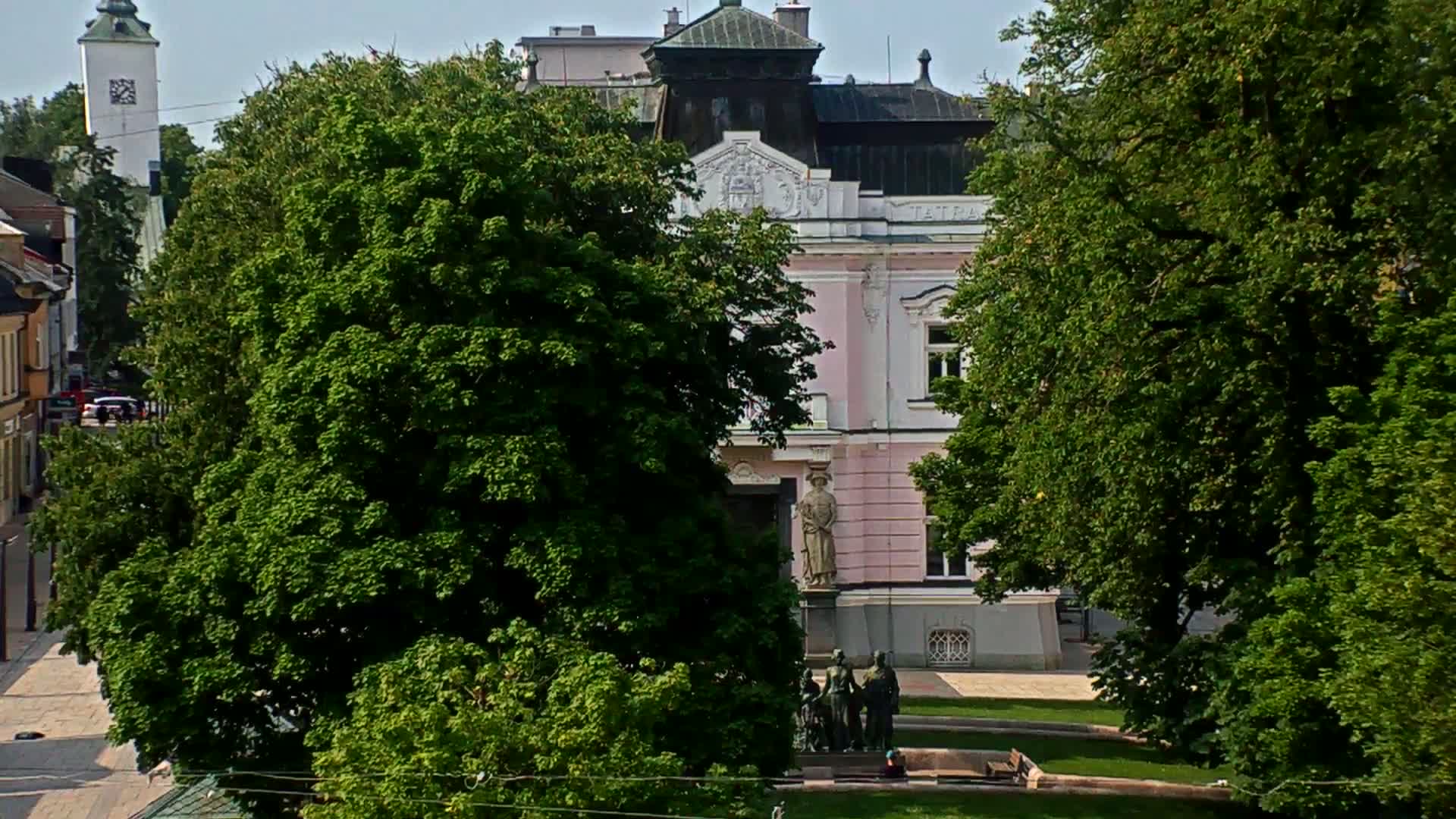 A pink building with a statue in front is partially obscured by lush green trees on a sunny day.