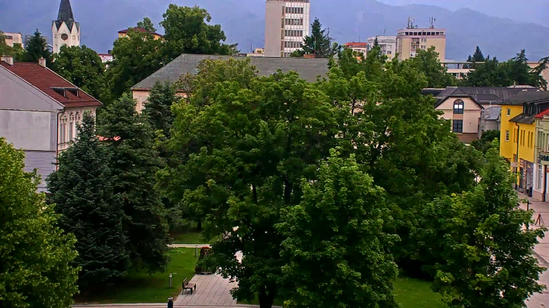 A town square with lush green trees, several buildings including a church steeple and a multi-story building, under an overcast sky.