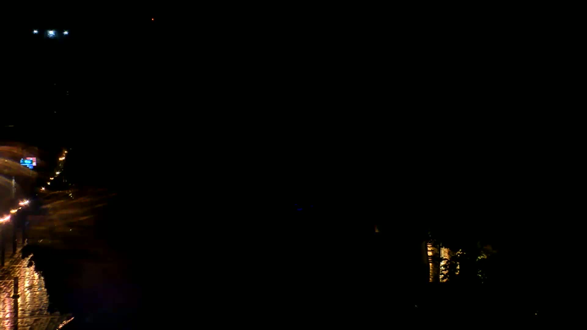 A nighttime view of a rain-slicked roadway with traffic lights and vehicle lights visible, and a dimly lit building in the background.