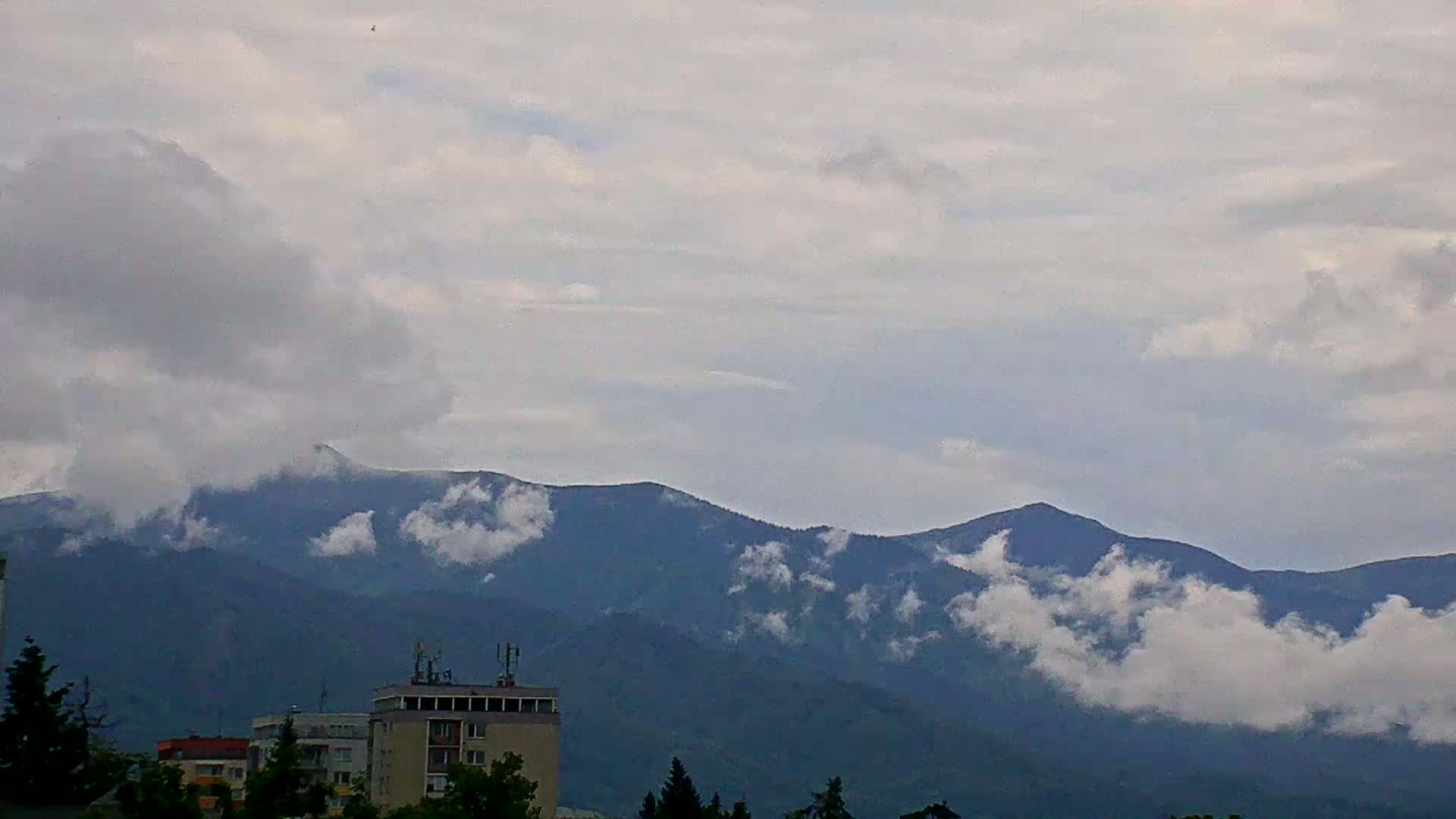 A partly cloudy day reveals a mountain range partially obscured by low-lying clouds, with a city skyline visible in the foreground.