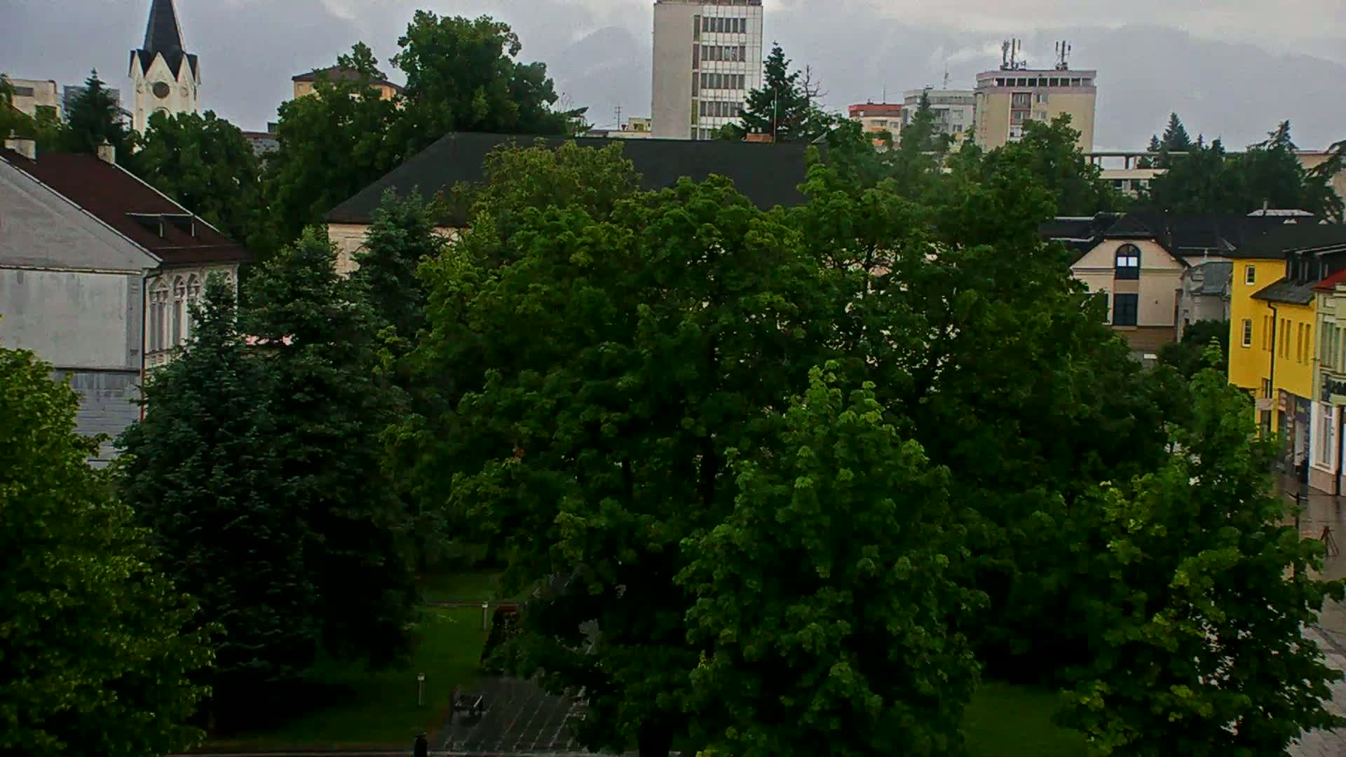 A town square with lush green trees, buildings of varying heights and colors, and an overcast sky.