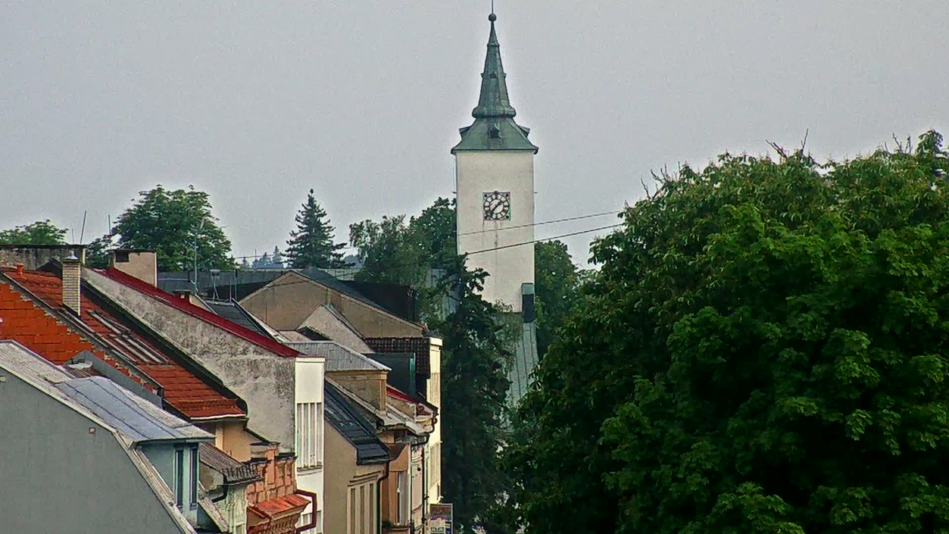 A white clock tower rises above a row of houses with red-tiled roofs, under an overcast sky.