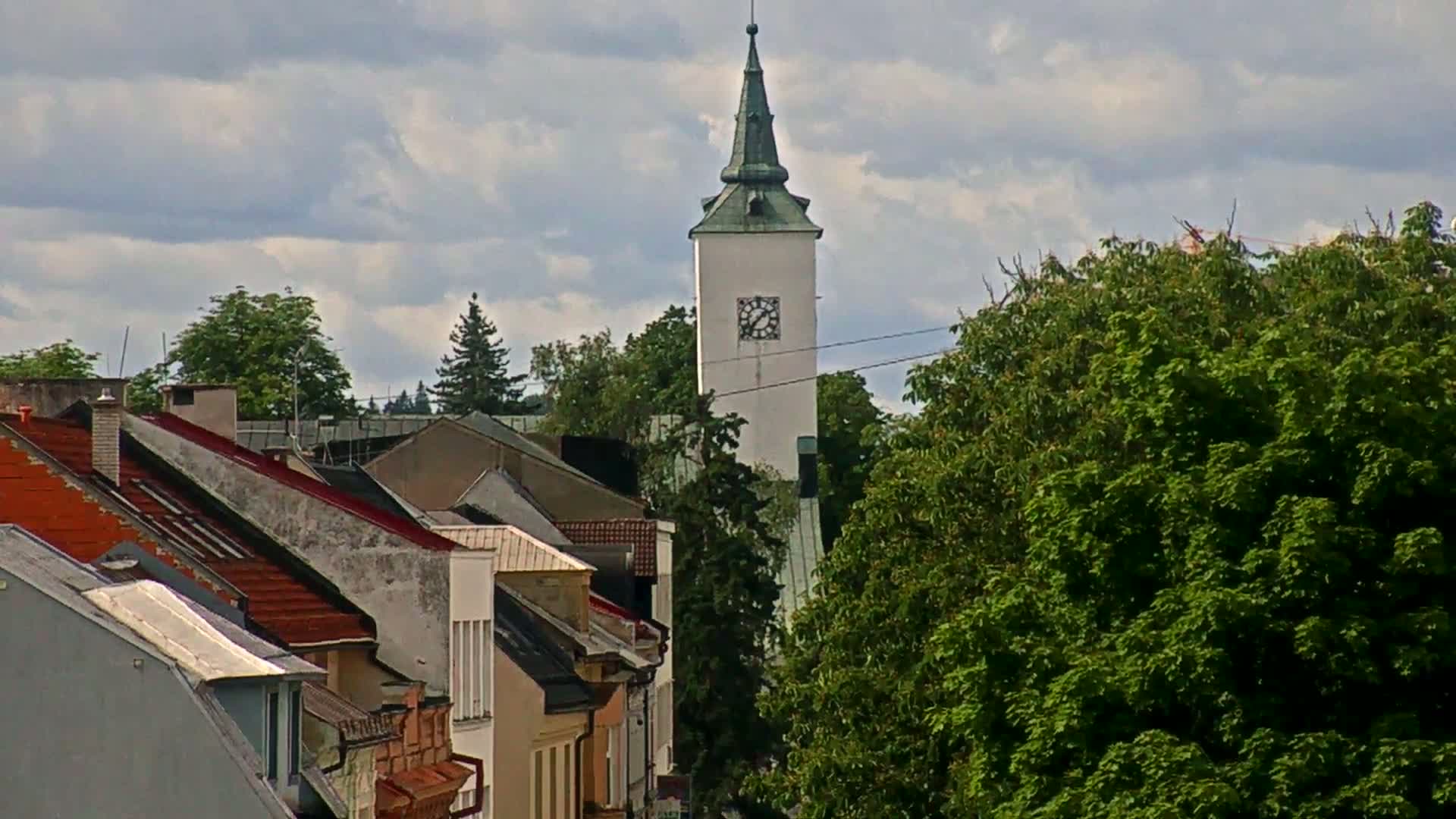 A white clock tower with a green spire rises above a row of houses under a partly cloudy sky.