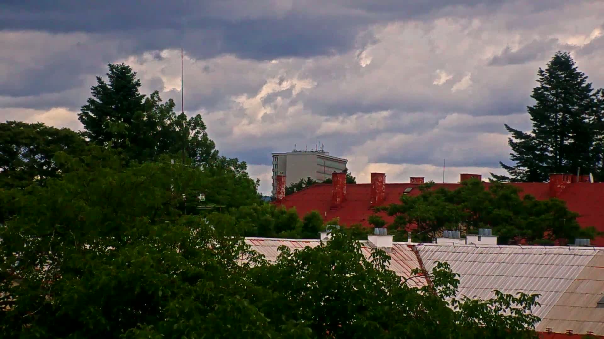 A view of rooftops with red and white tiles, trees, and a multi-story building in the background under a cloudy sky.
