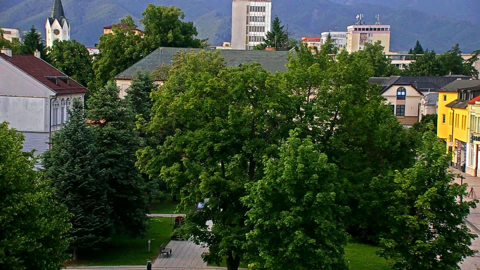 Lush green trees fill a town square, with various buildings including a church steeple and multi-story structures visible in the background under a mostly clear sky.