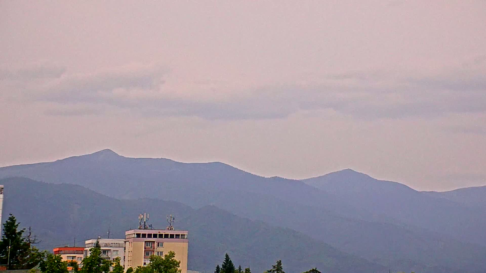 A partly cloudy sky overlooks a cityscape with several buildings and trees in the foreground, and a range of mountains in the background.