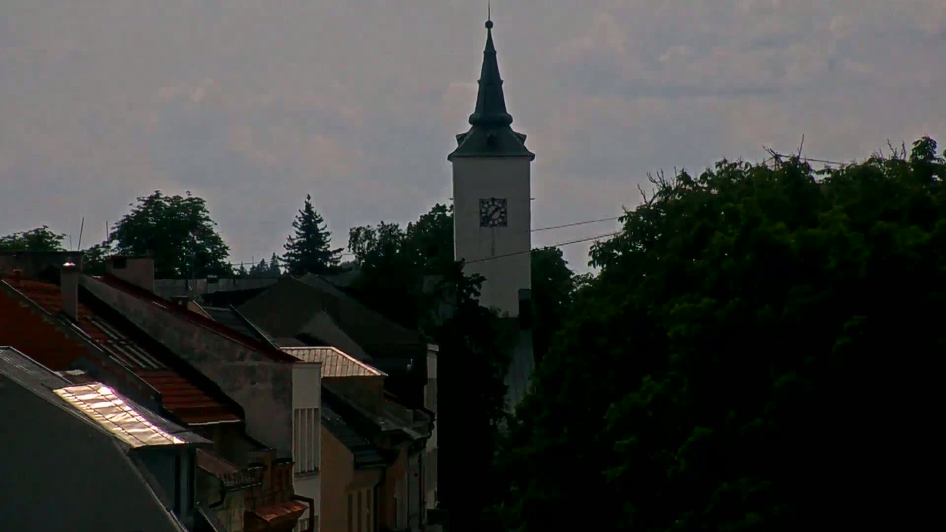 A white clock tower with a dark green steeple rises above a cluster of houses and lush green trees under a cloudy sky.