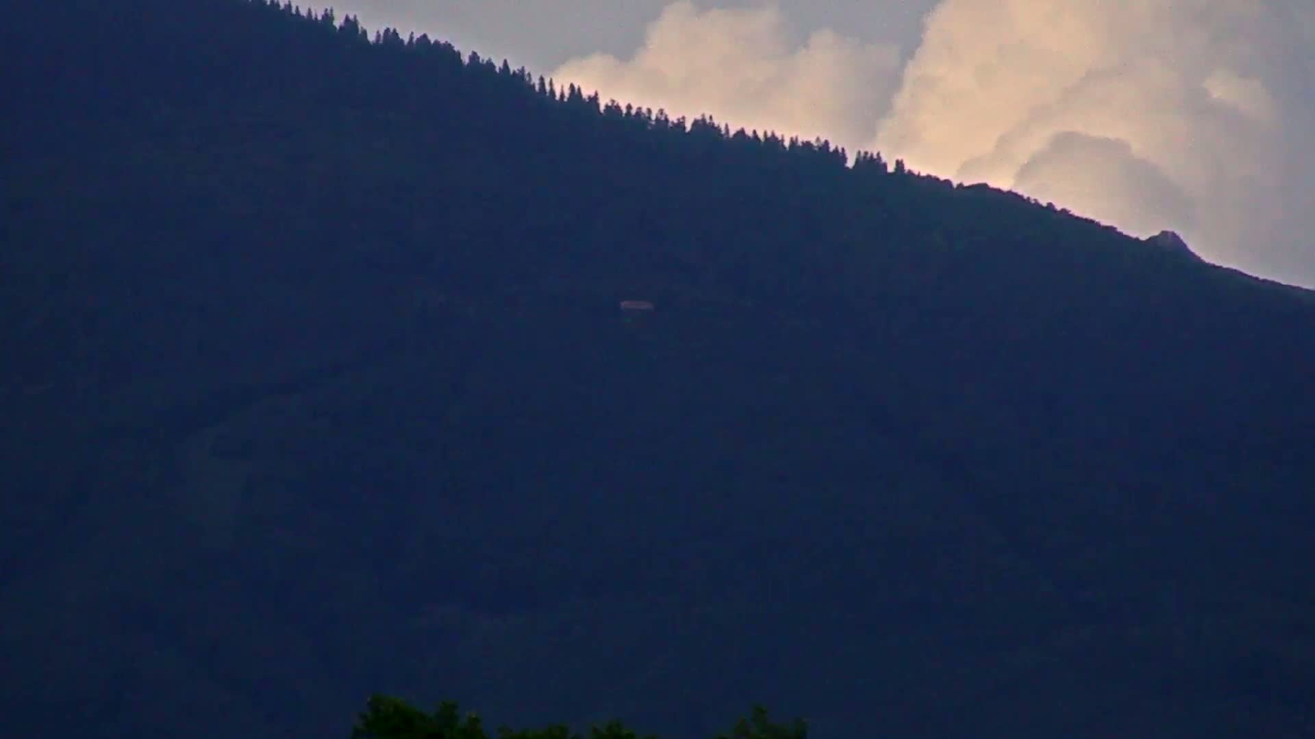 A dark, forested mountainside is partially illuminated by a partly cloudy sky.