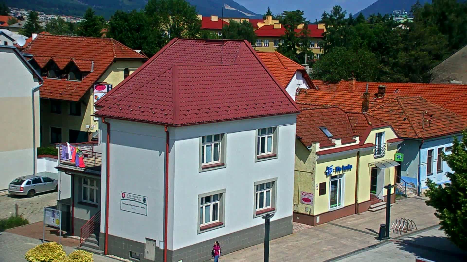 A light-grey building with a red roof stands prominently among other buildings with red-tiled roofs, all under a sunny sky with mountains in the background.