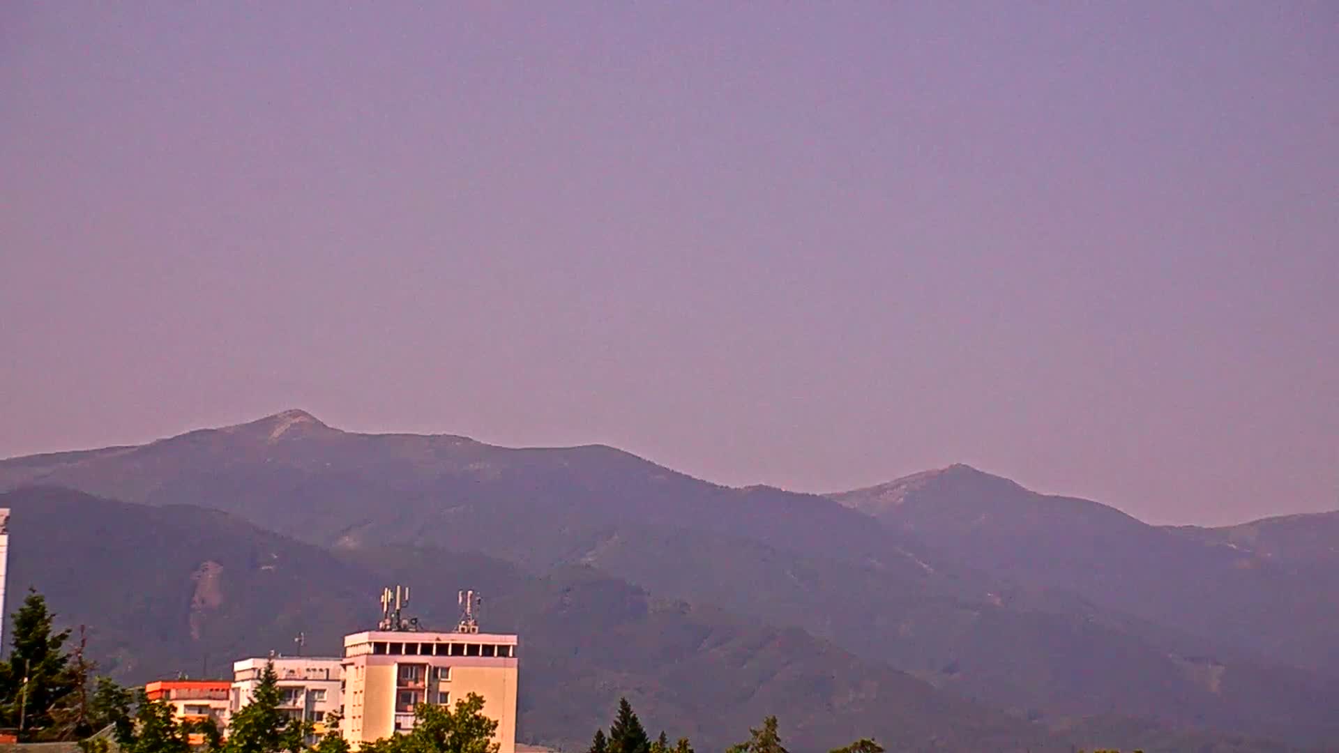 Under a hazy purple sky, a range of dark purple mountains rises behind a group of buildings, some of which are multi-story apartments.
