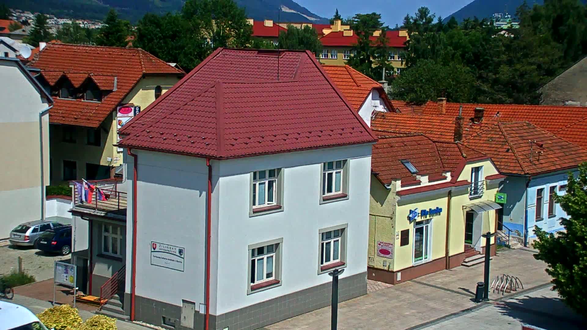 A light-grey building with a red roof stands among other buildings with red tile roofs, under a sunny sky and against a backdrop of mountains.