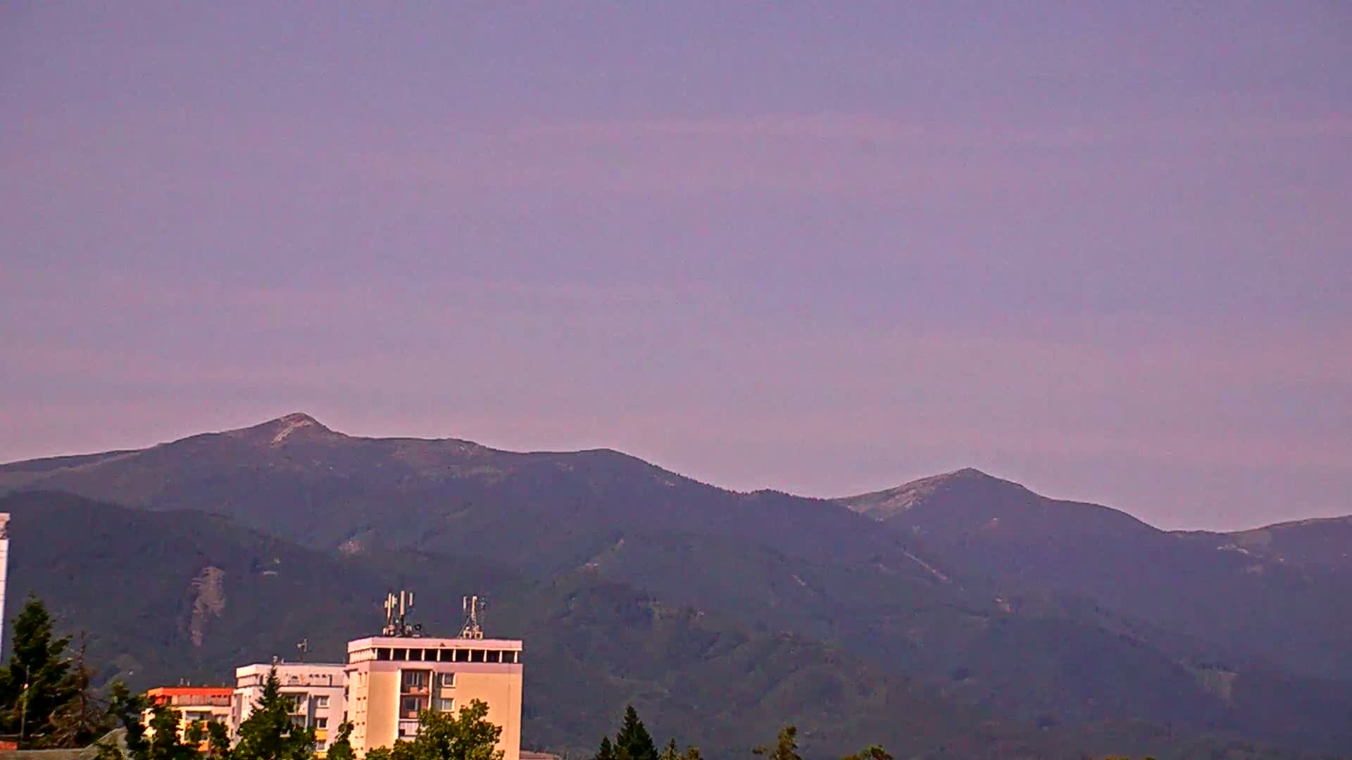 Under a pale purple sky, a range of dark green mountains rises behind a group of buildings.