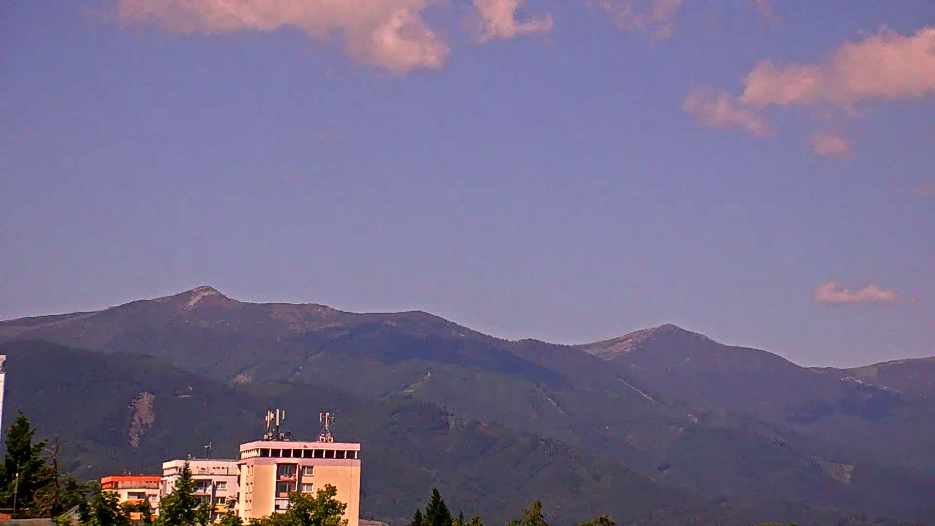 A partly cloudy sky overlooks a range of dark green mountains behind a city skyline featuring several buildings.