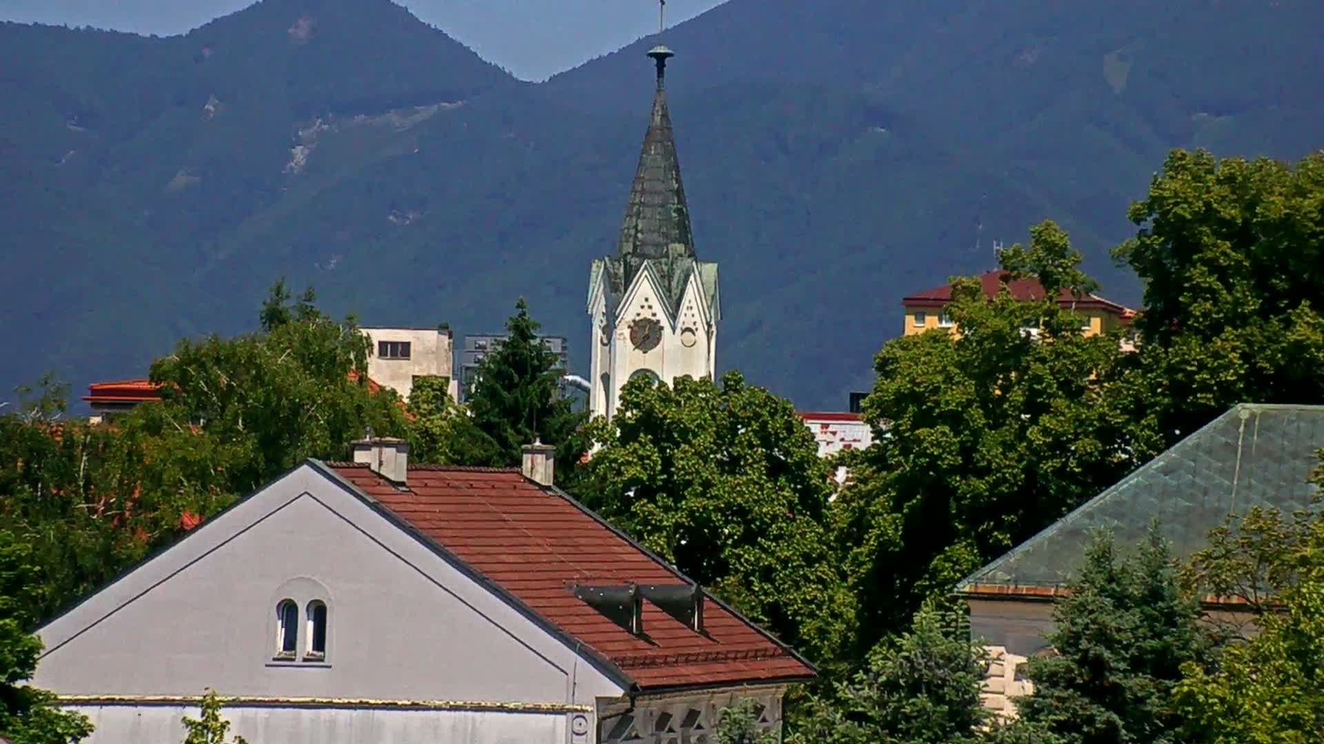 A tall church steeple is visible behind a cluster of houses and leafy green trees, all set against a backdrop of distant, blue-tinged mountains under a clear sky.