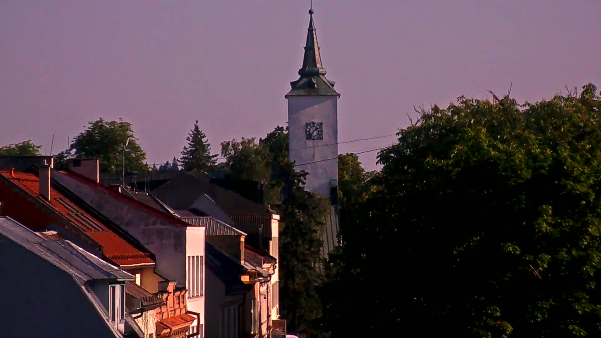A tall, white clock tower with a green spire rises above a row of houses with red tile roofs under a clear, pale sky.