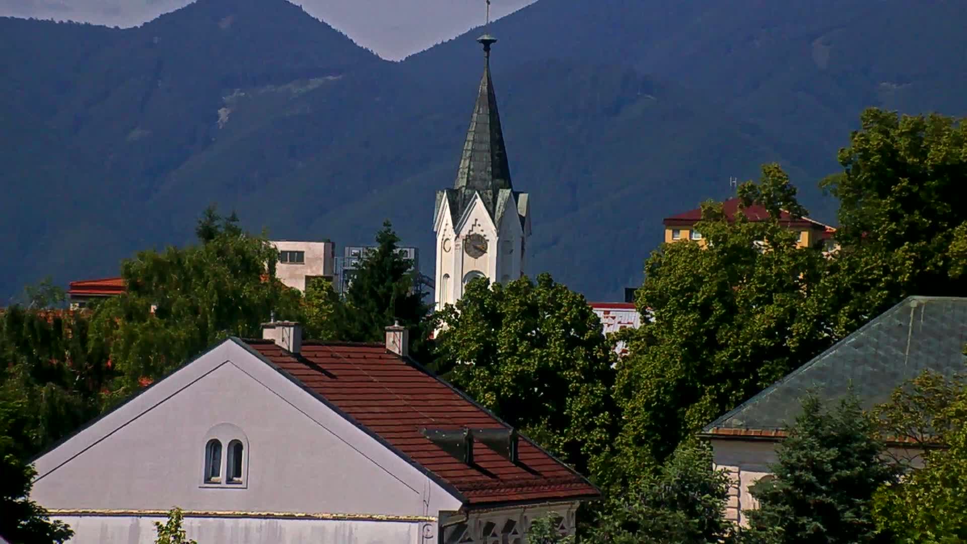 A white church steeple with a clock is partially visible behind several buildings and lush green trees, all set against a backdrop of dark blue mountains under a mostly clear sky.