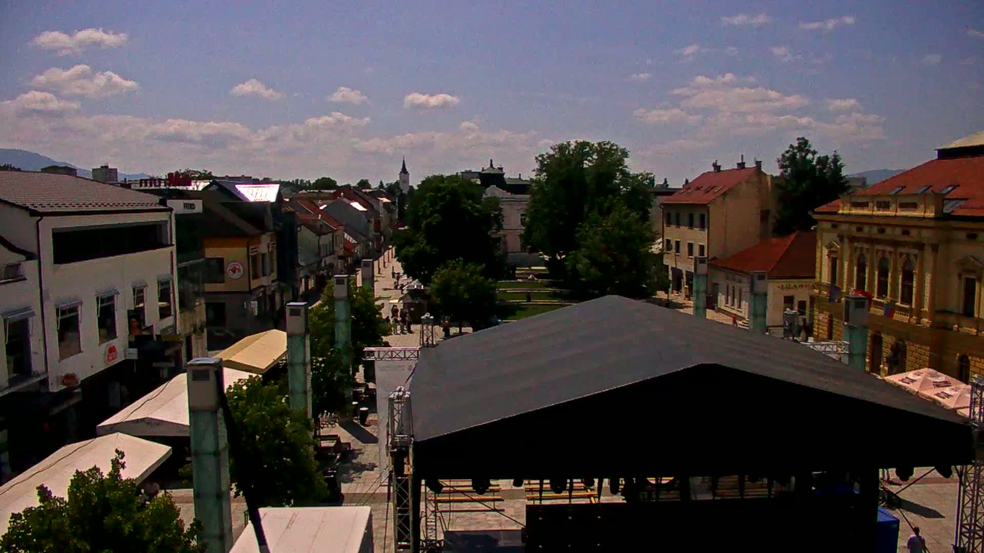 A sunny day reveals a town square with buildings, trees, a stage, and people; mountains are visible in the background.