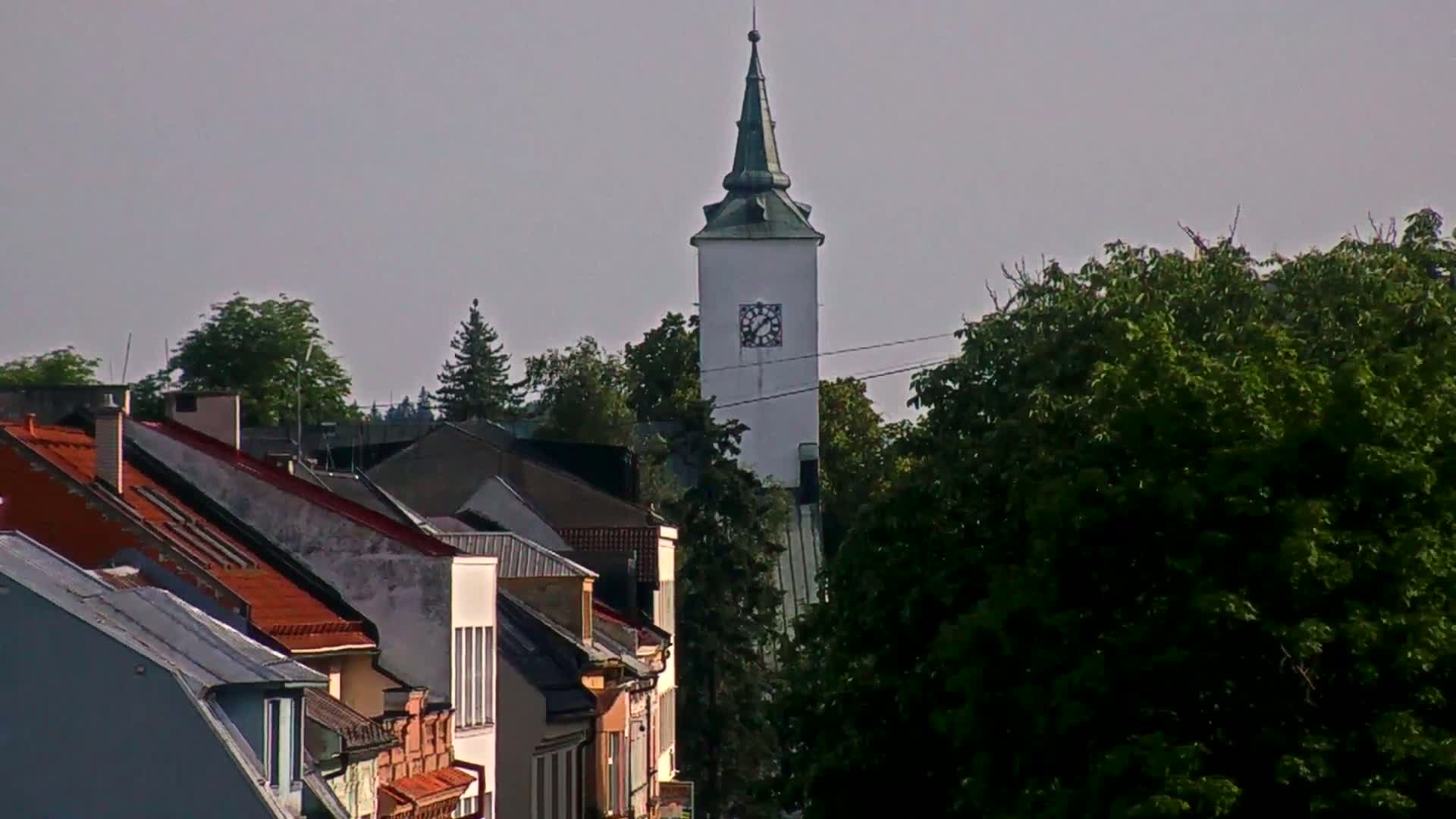 A white clock tower rises above a row of buildings with red-tiled roofs, under a mostly cloudy sky.