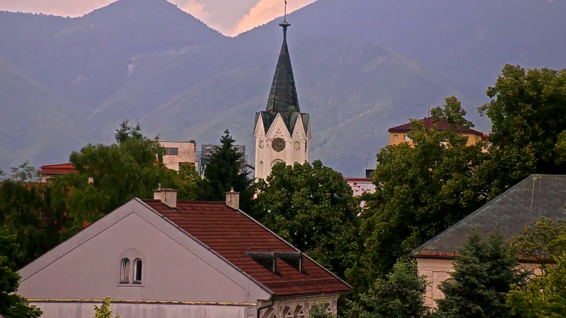 A tall church steeple rises above a collection of houses and lush green trees, all set against a backdrop of hazy mountains under a cloudy sky.