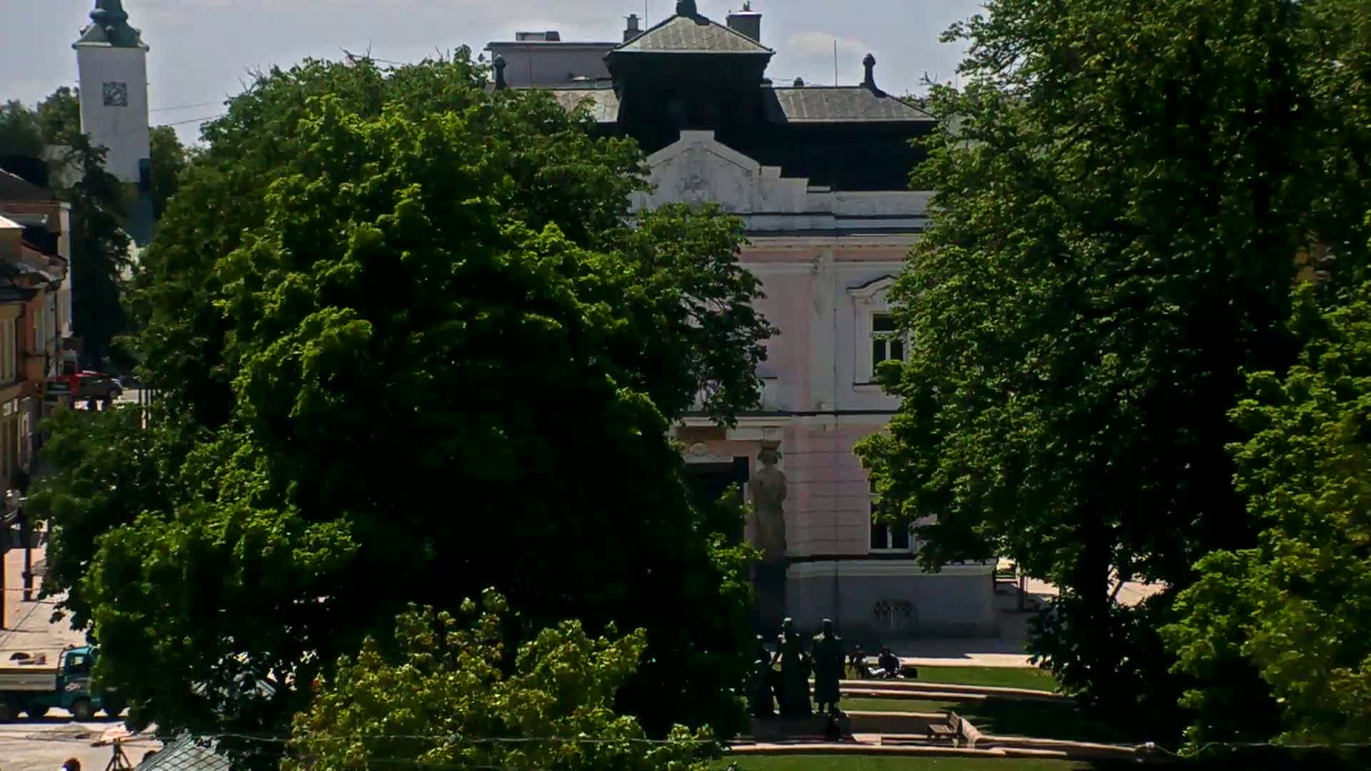 A pink building partially obscured by lush green trees, under a sunny sky, with a group of statues in a plaza.
