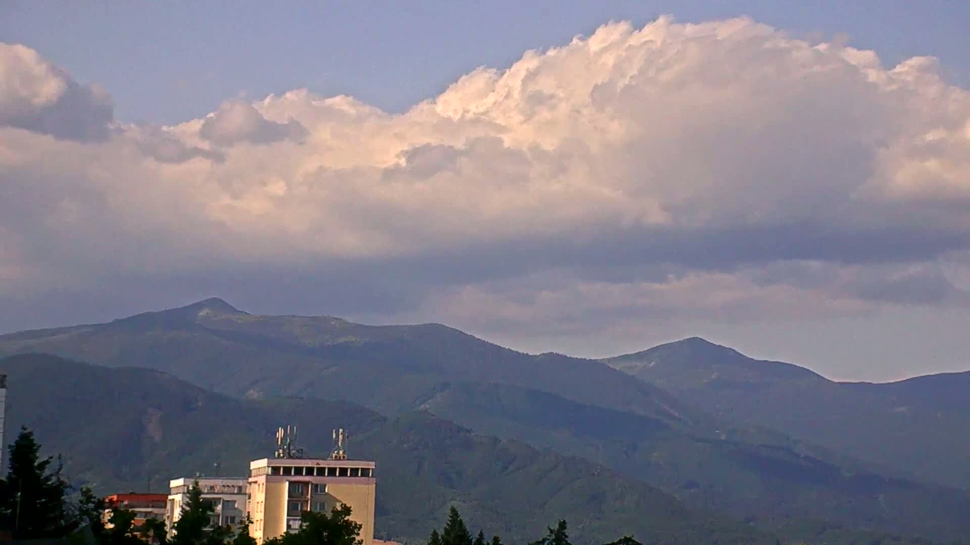 A large, fluffy cloud hangs over a range of dark green mountains, partially obscuring them from view, with several buildings and trees in the foreground.