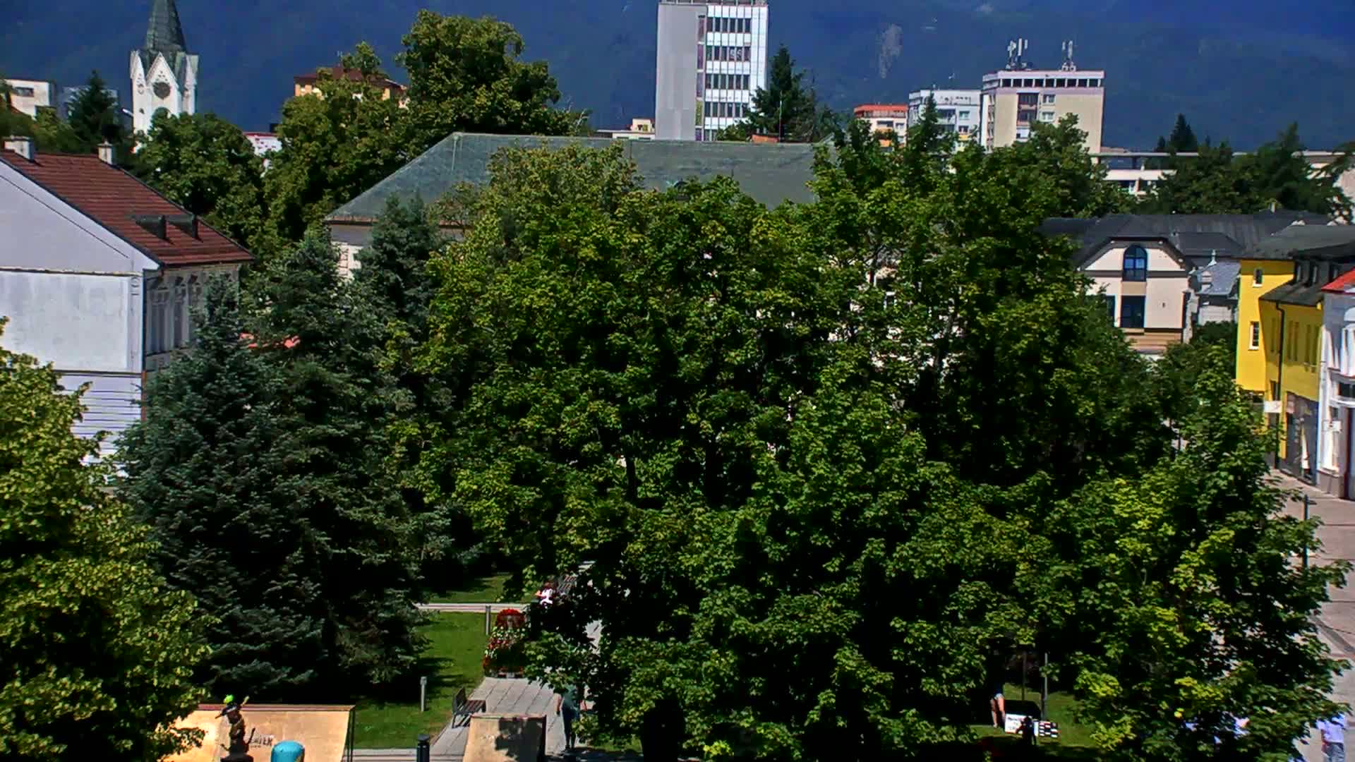 A town square scene on a sunny day, featuring lush green trees surrounding buildings of varying heights and architectural styles, including a church steeple in the background.
