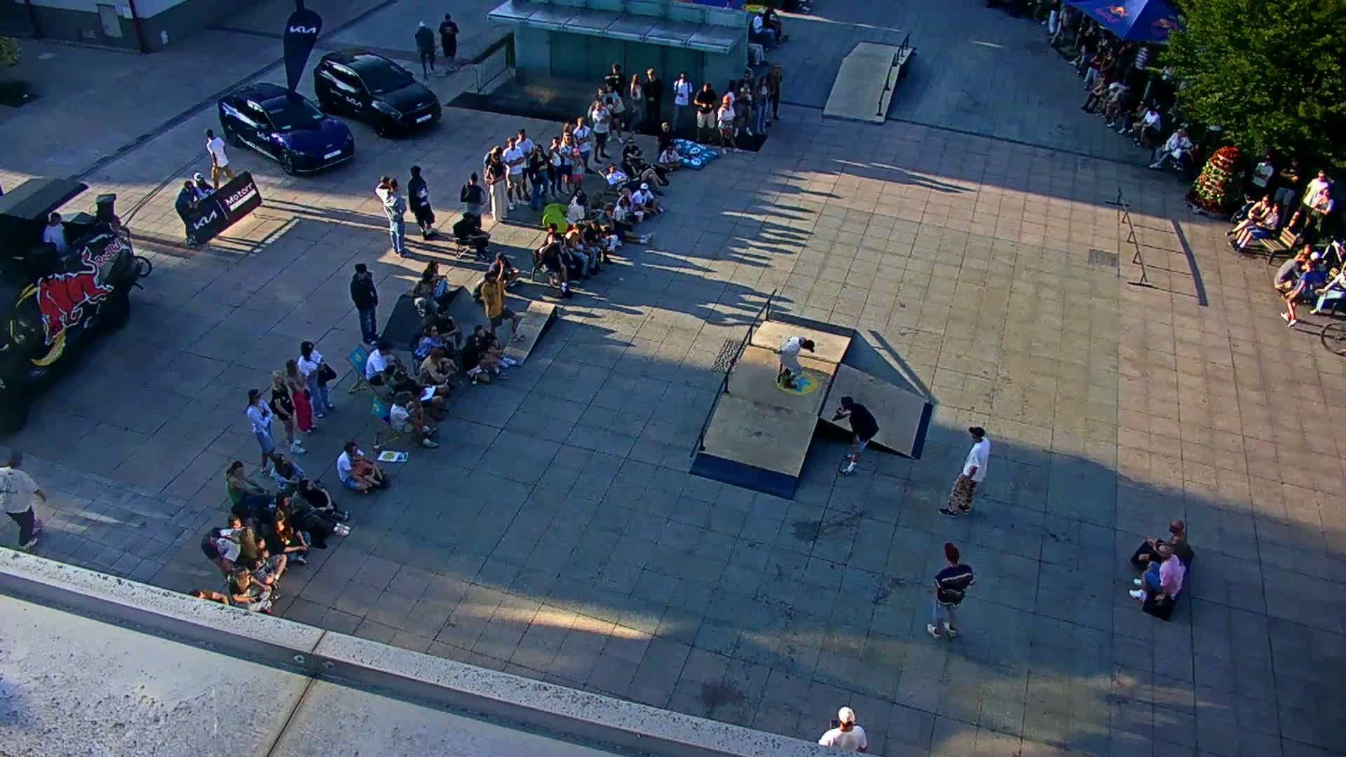 A skateboarding event takes place outdoors on a sunny day in a paved plaza, with spectators seated on the ground watching a skateboarder perform tricks on a ramp.