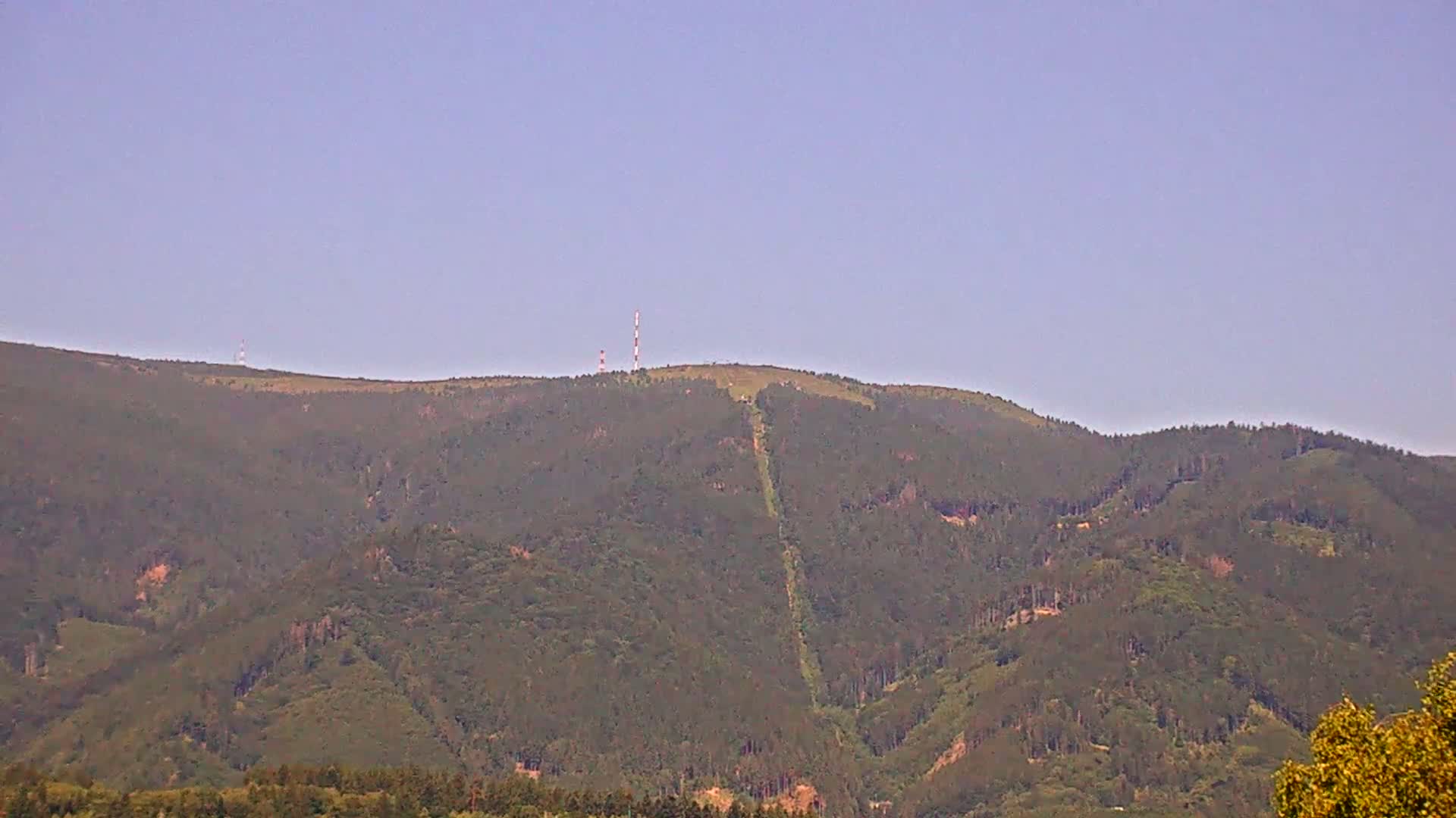 A distant, forested mountain ridge with two communication towers on its peak is visible under a clear, light-blue sky.