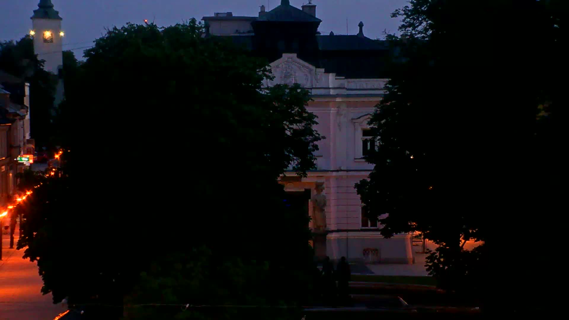 A light pink building with a statue stands at dusk, partially obscured by trees, with a clock tower visible in the background and streetlights illuminating a street.